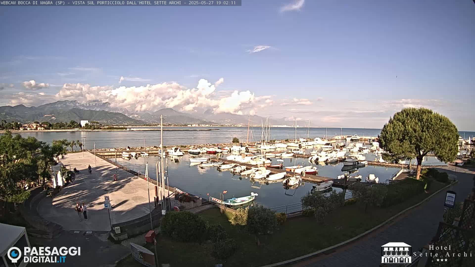 A partly cloudy day shows a marina filled with boats, a paved area with a few people, and mountains in the distance.