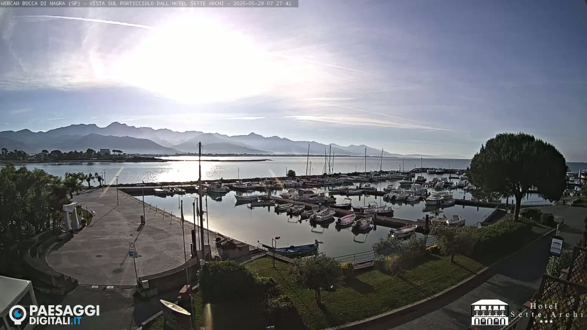 A marina filled with numerous boats is situated by a calm body of water, with mountains visible in the distance under a mostly clear, bright sky.