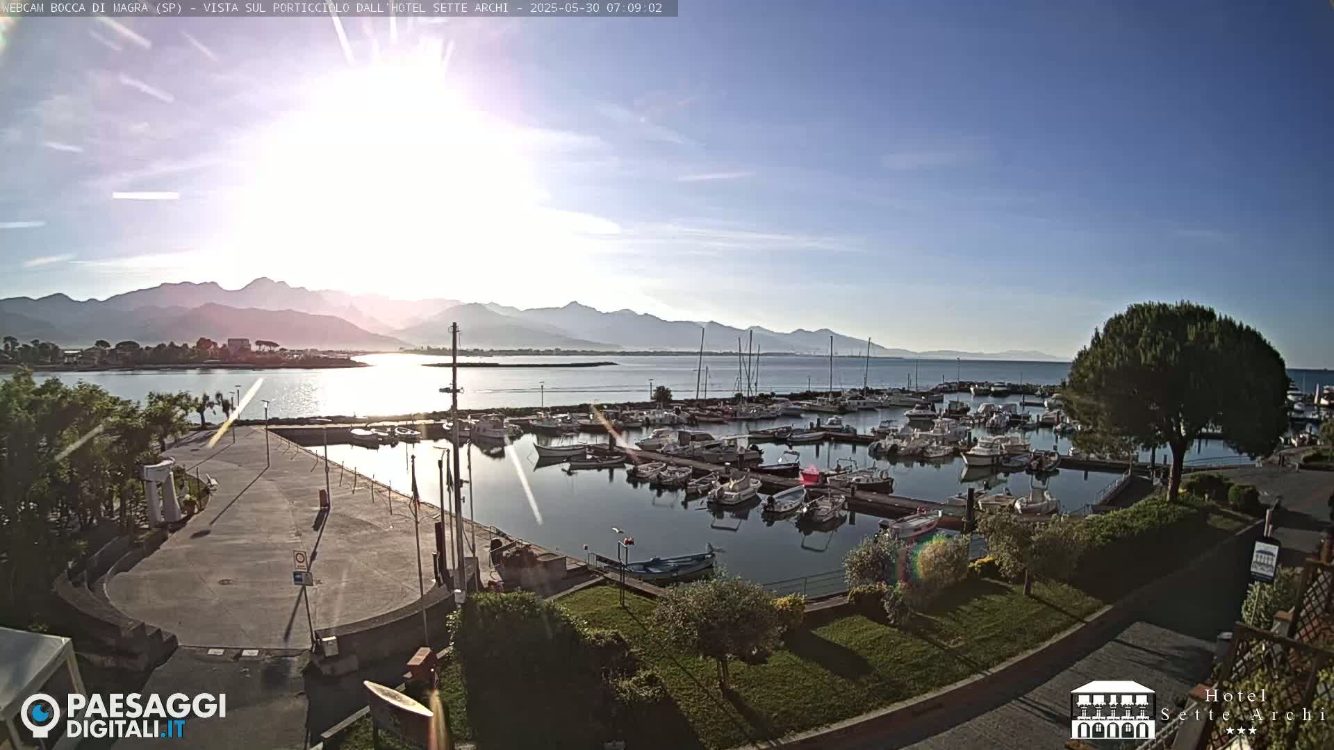 A marina filled with numerous boats is situated next to a paved area, overlooking calm water and distant mountains under a mostly clear, sunny sky.