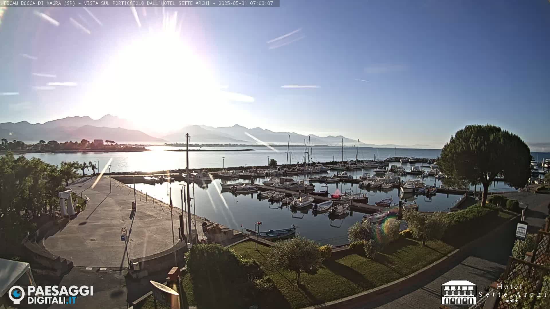 A marina filled with boats is situated near a calm body of water under a clear, sunny sky, with mountains visible in the distance.