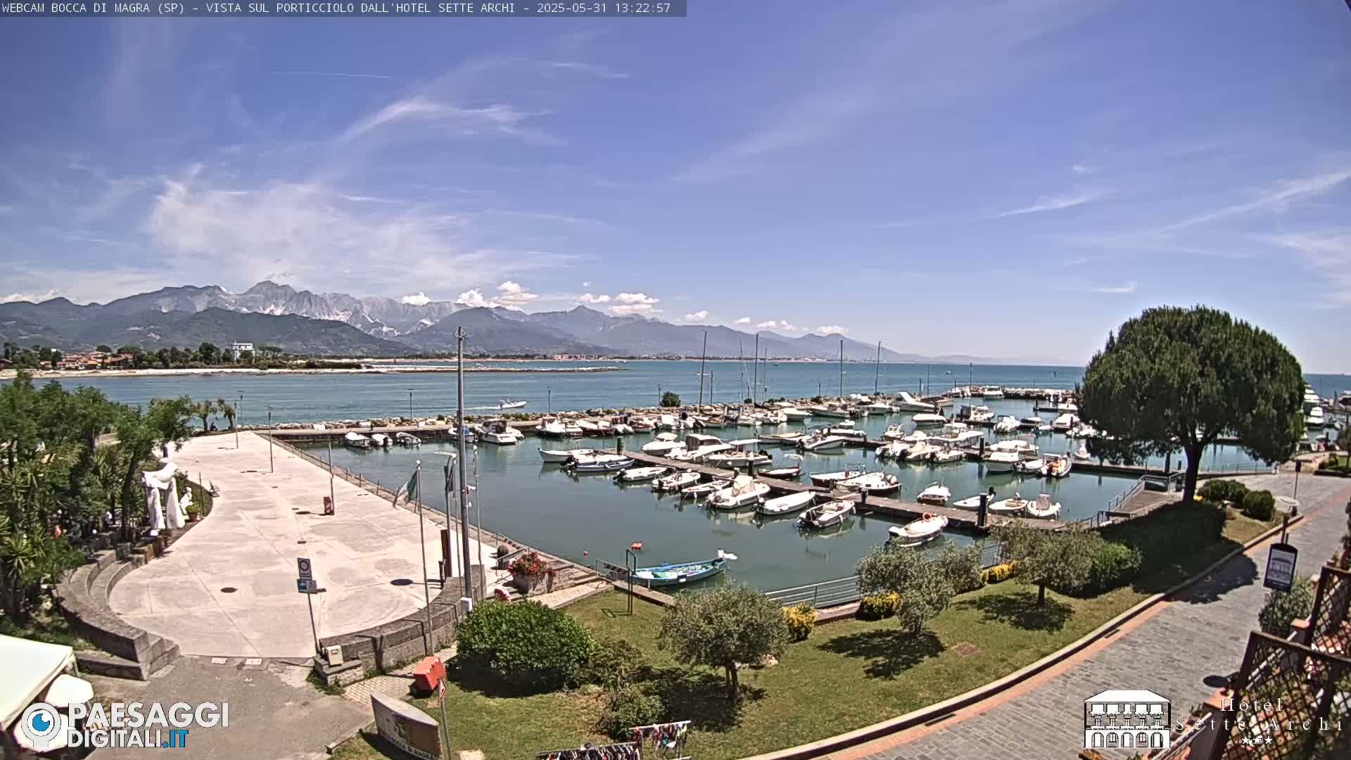 A marina filled with boats is situated on calm water under a mostly sunny sky, with mountains visible in the distance.