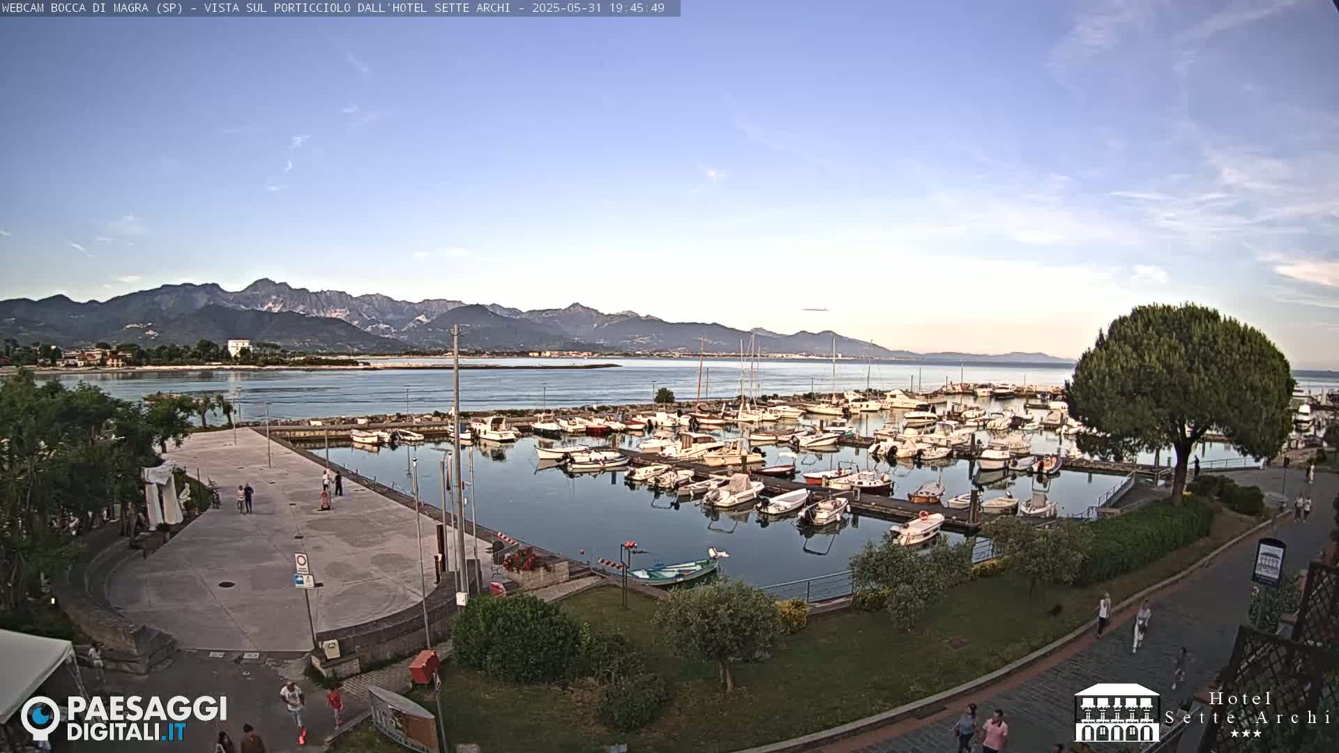 A harbor filled with numerous boats is situated next to a paved area with people walking, all under a clear, evening sky with mountains in the distance.