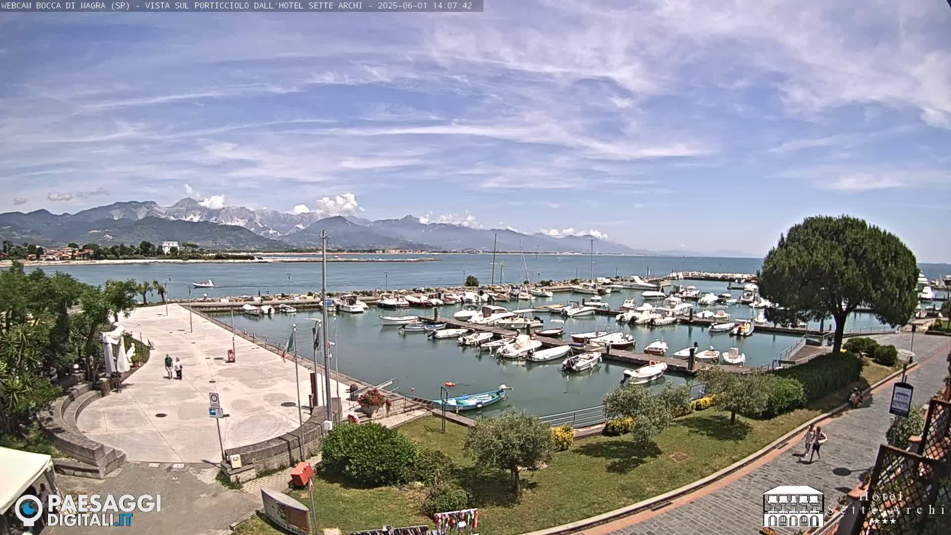 A marina filled with boats is situated beside a paved plaza, overlooking a calm body of water and mountains under a mostly sunny sky.