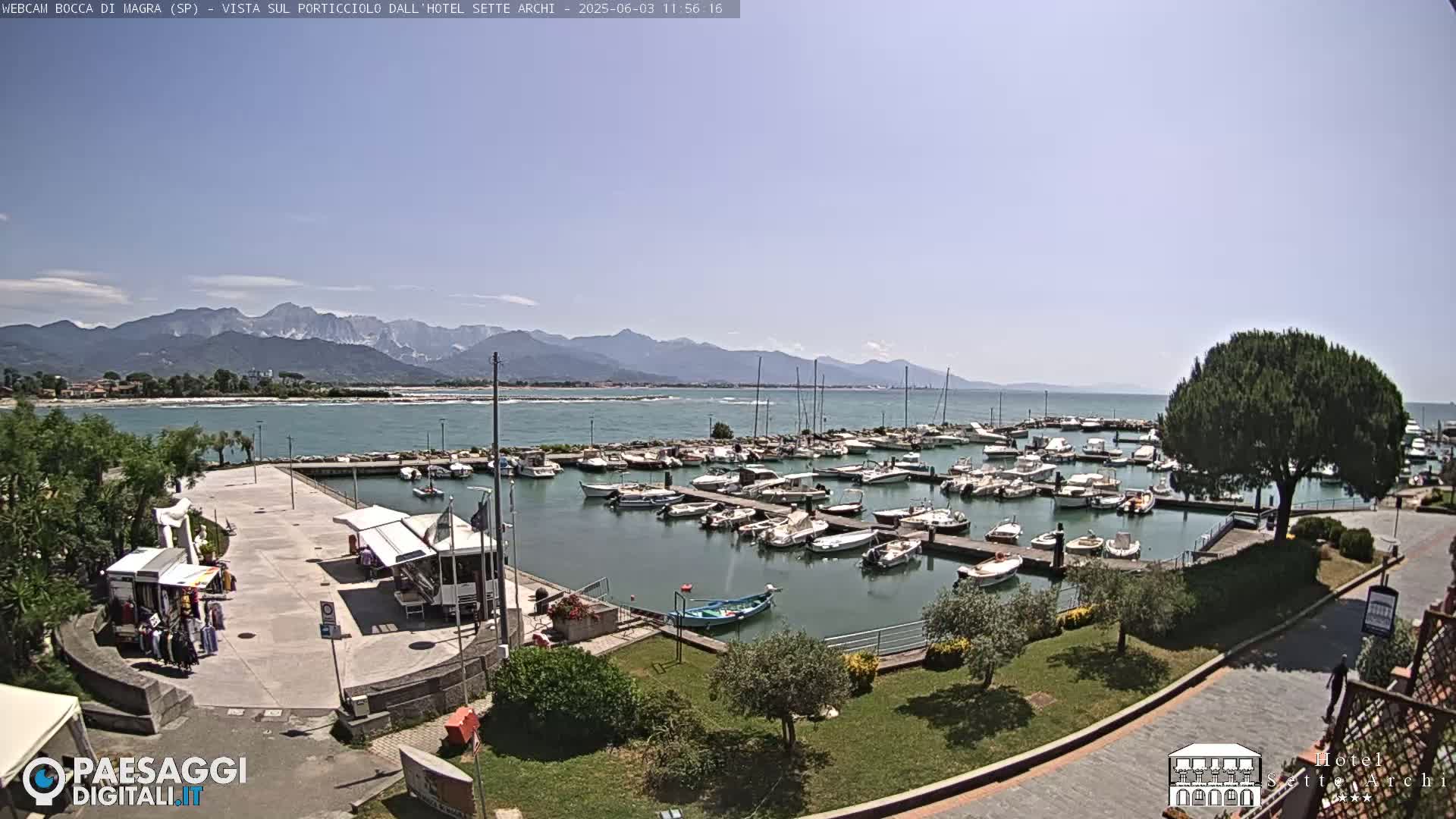 A marina filled with numerous boats is situated near a shoreline, with mountains visible in the background under a clear, sunny sky.