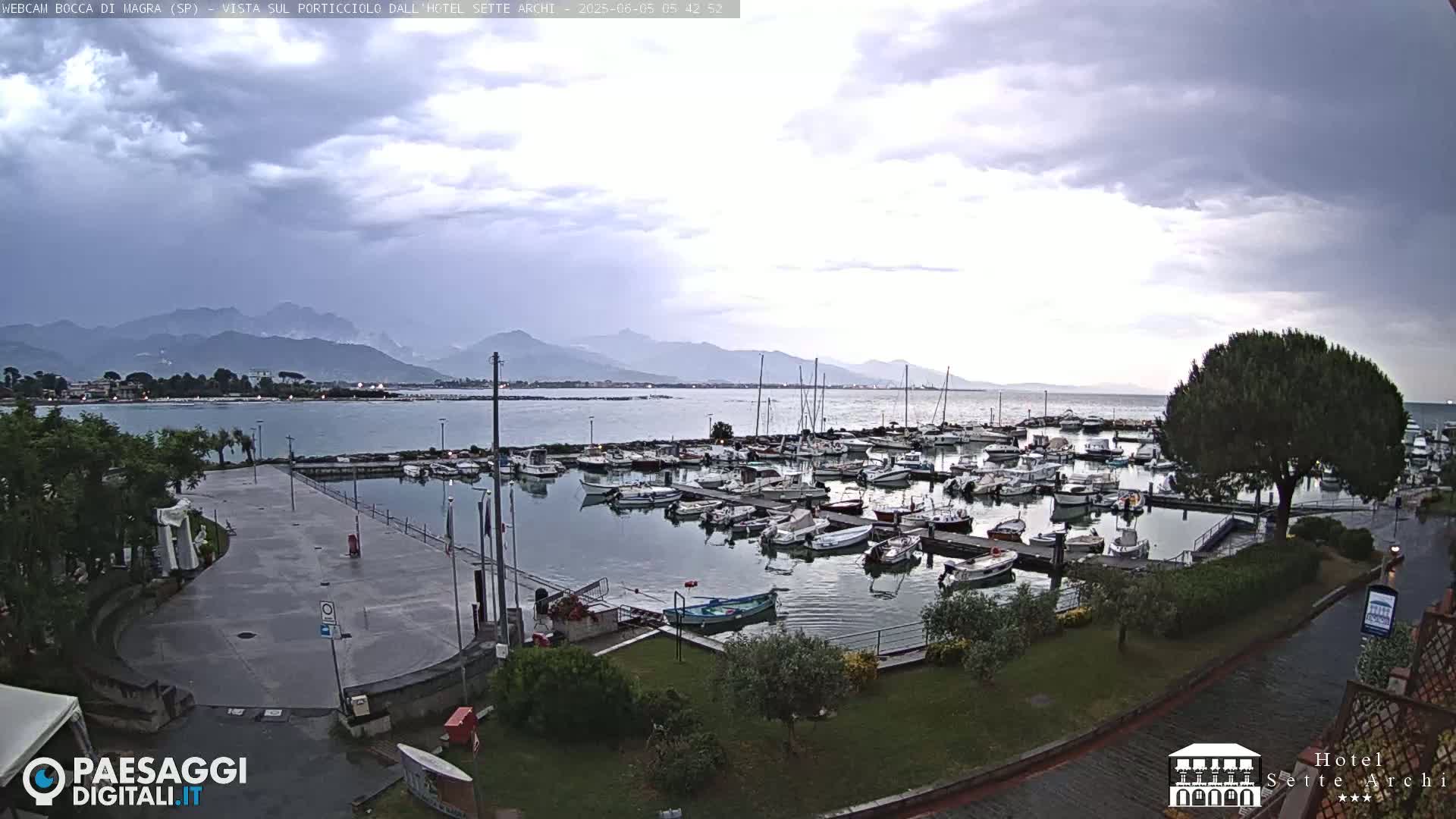 A harbor filled with numerous boats is situated near a paved area, with mountains visible in the distance under a cloudy sky.