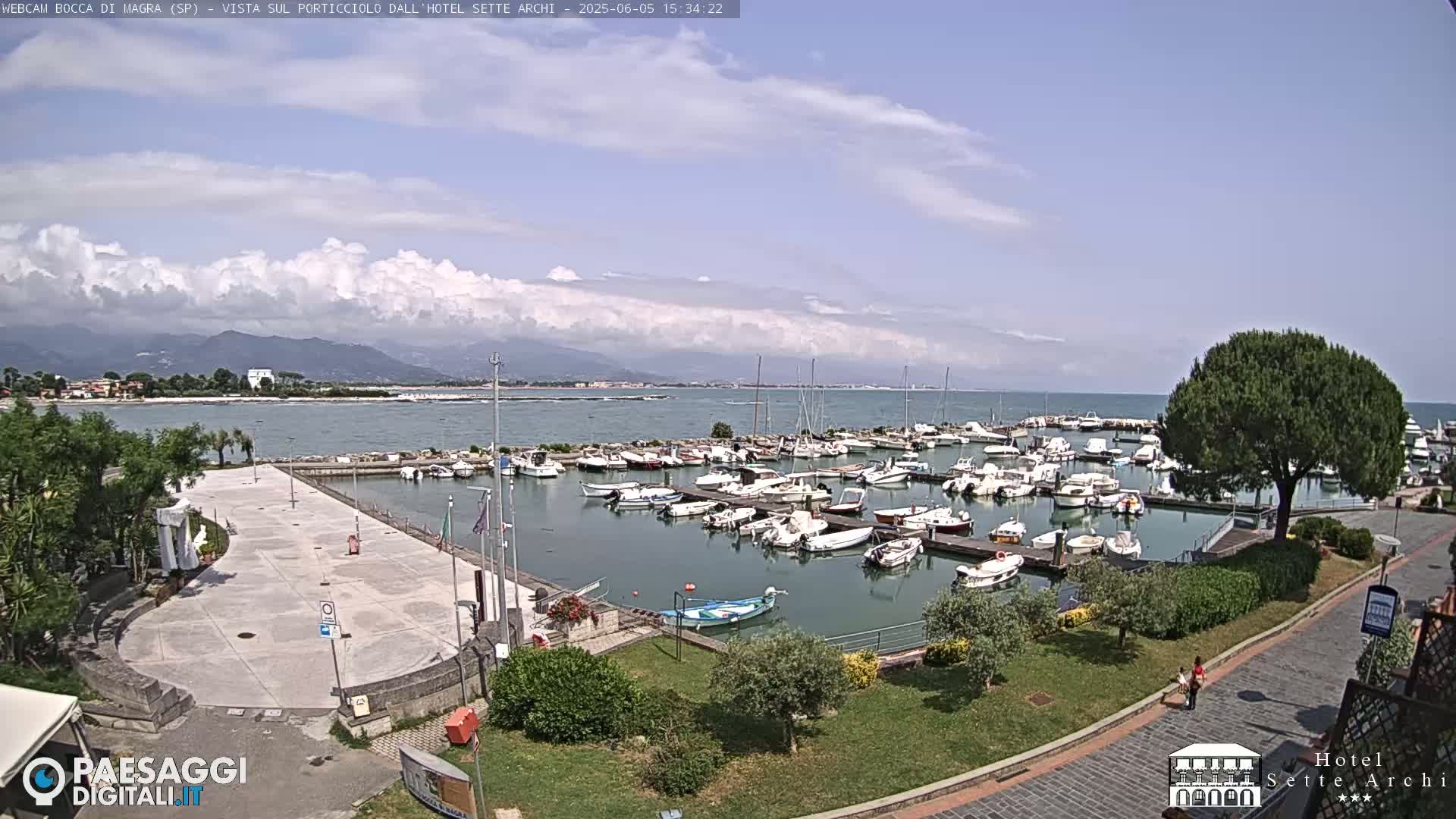A harbor filled with boats is situated next to a paved plaza and grassy area, all overlooking a calm sea under a partly cloudy sky with mountains in the distance.
