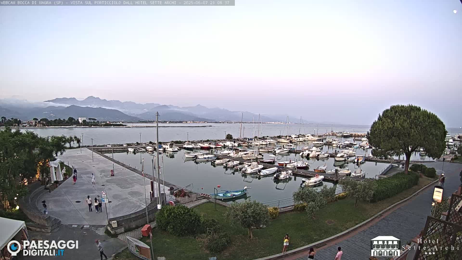 A calm harbor filled with numerous boats is situated near a paved plaza with several people walking, all under a twilight sky with distant mountains visible in the background.
