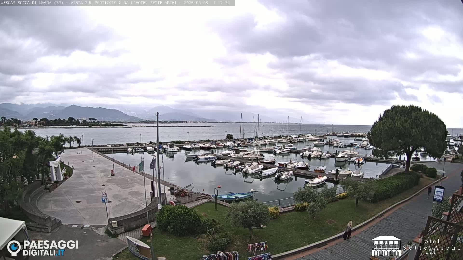 A harbor filled with numerous boats is visible under a cloudy sky, with mountains in the distance.