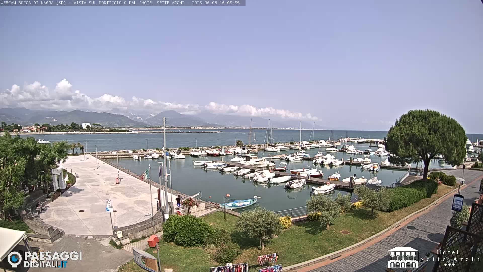A partly sunny day overlooks a harbor filled with numerous boats, a paved plaza, and green landscaping, with mountains visible in the distance.
