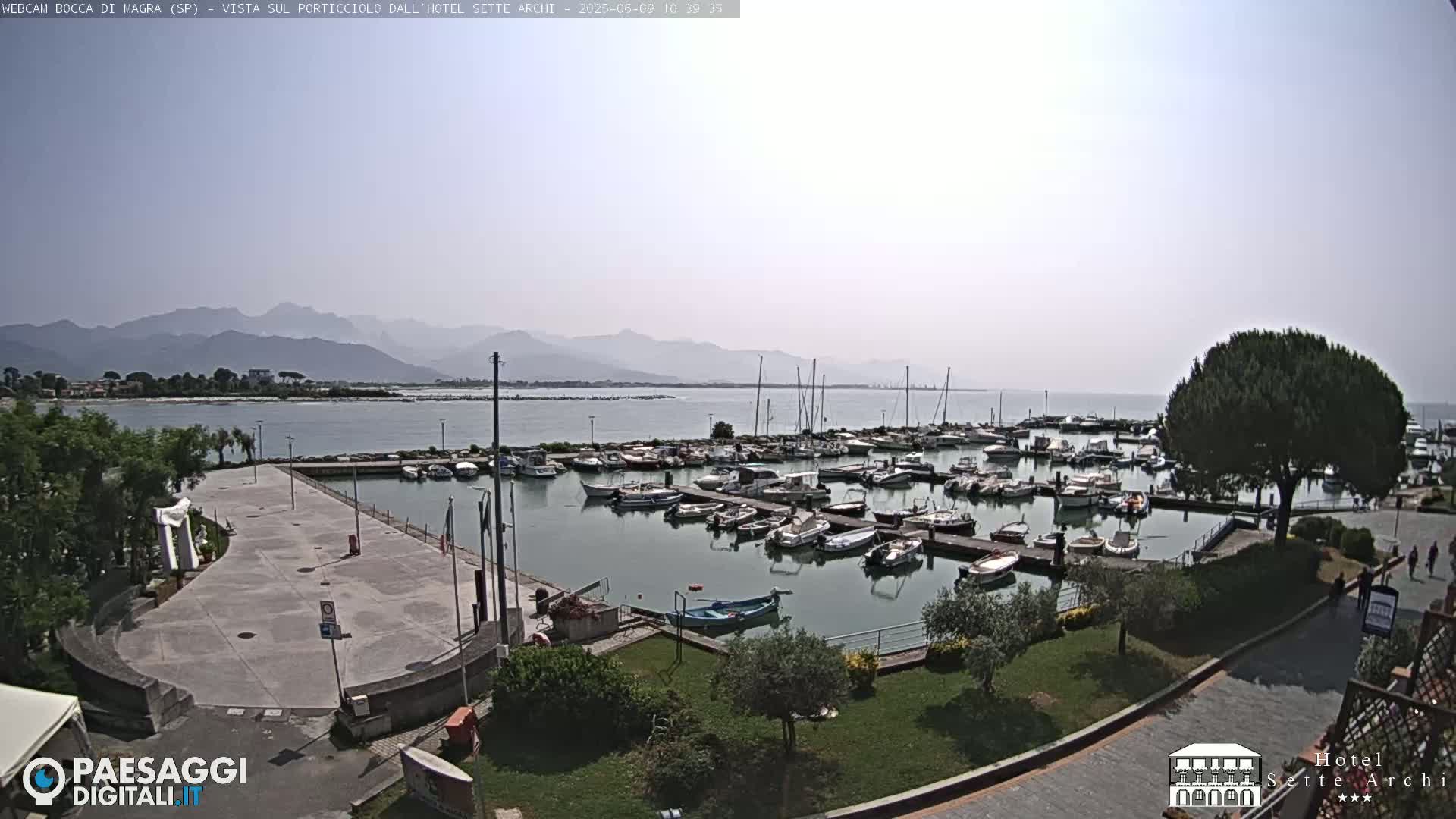 A marina filled with numerous boats is situated beside a paved plaza, overlooking calm water and distant mountains under a hazy sky.