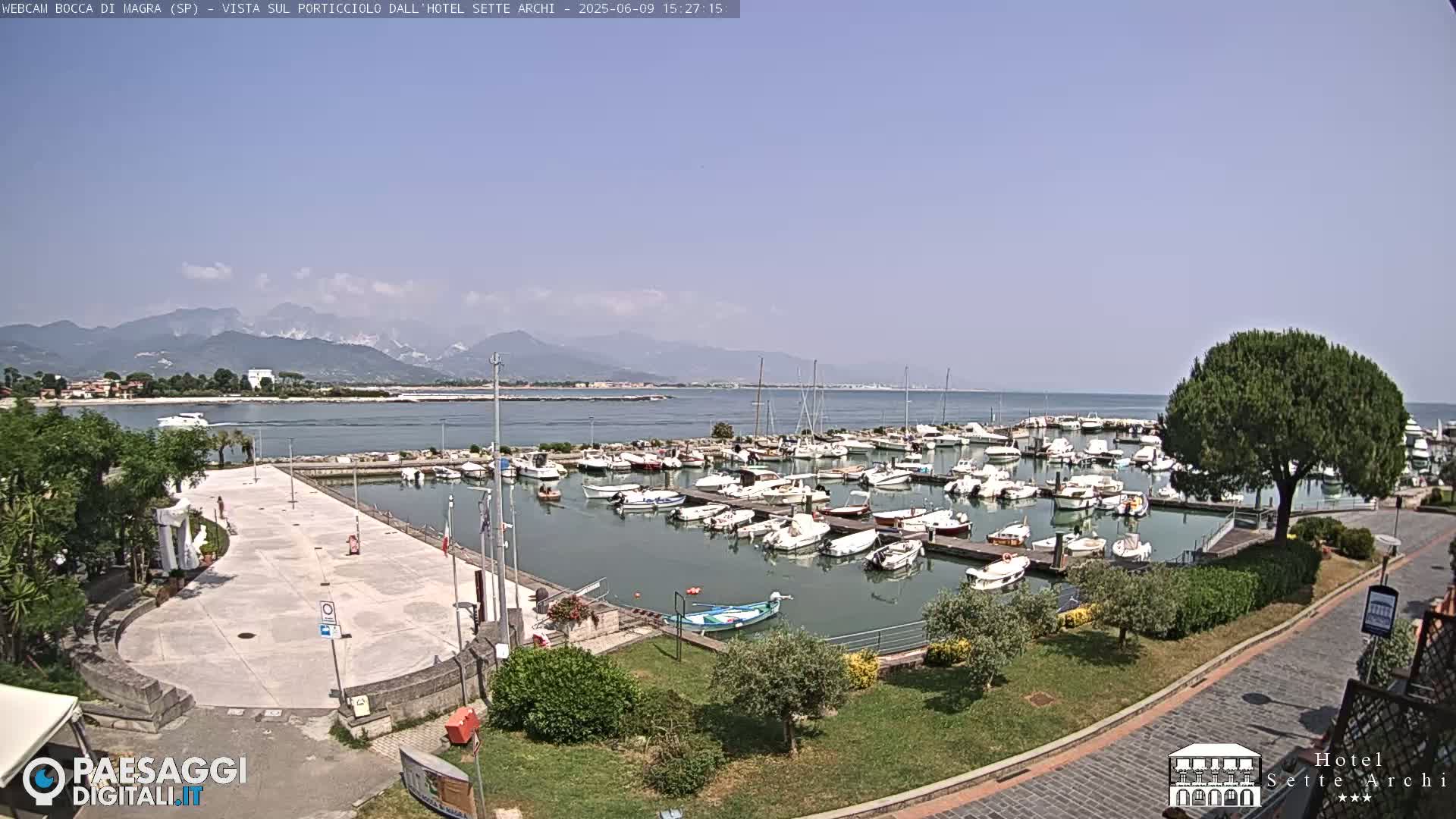 A marina filled with numerous boats is situated next to a paved plaza, overlooking calm water and distant mountains under a clear sky.
