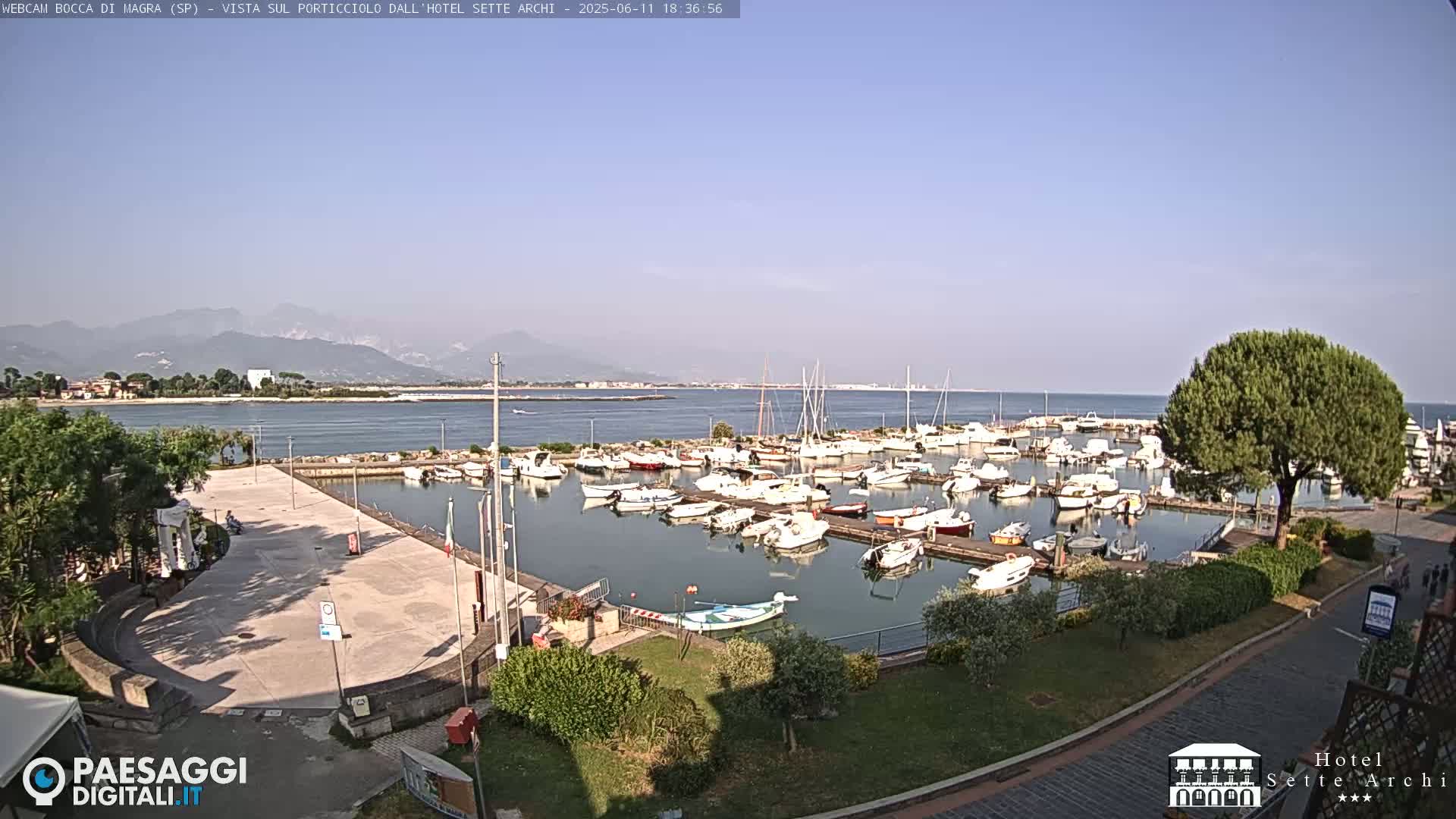 A marina filled with numerous boats is situated near a paved plaza, with mountains visible in the distance under a clear, sunny sky.