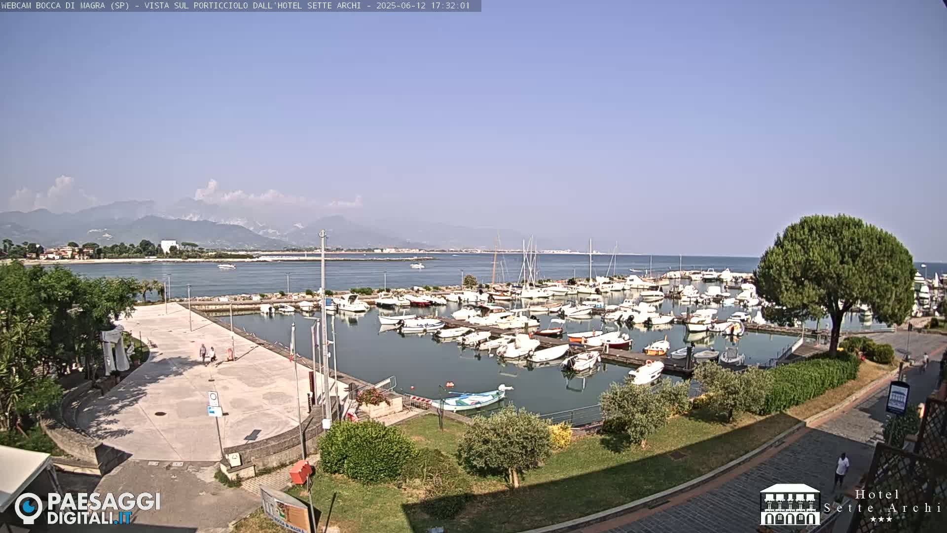A marina filled with numerous boats is situated near a paved plaza, with mountains visible in the distance under a clear, sunny sky.