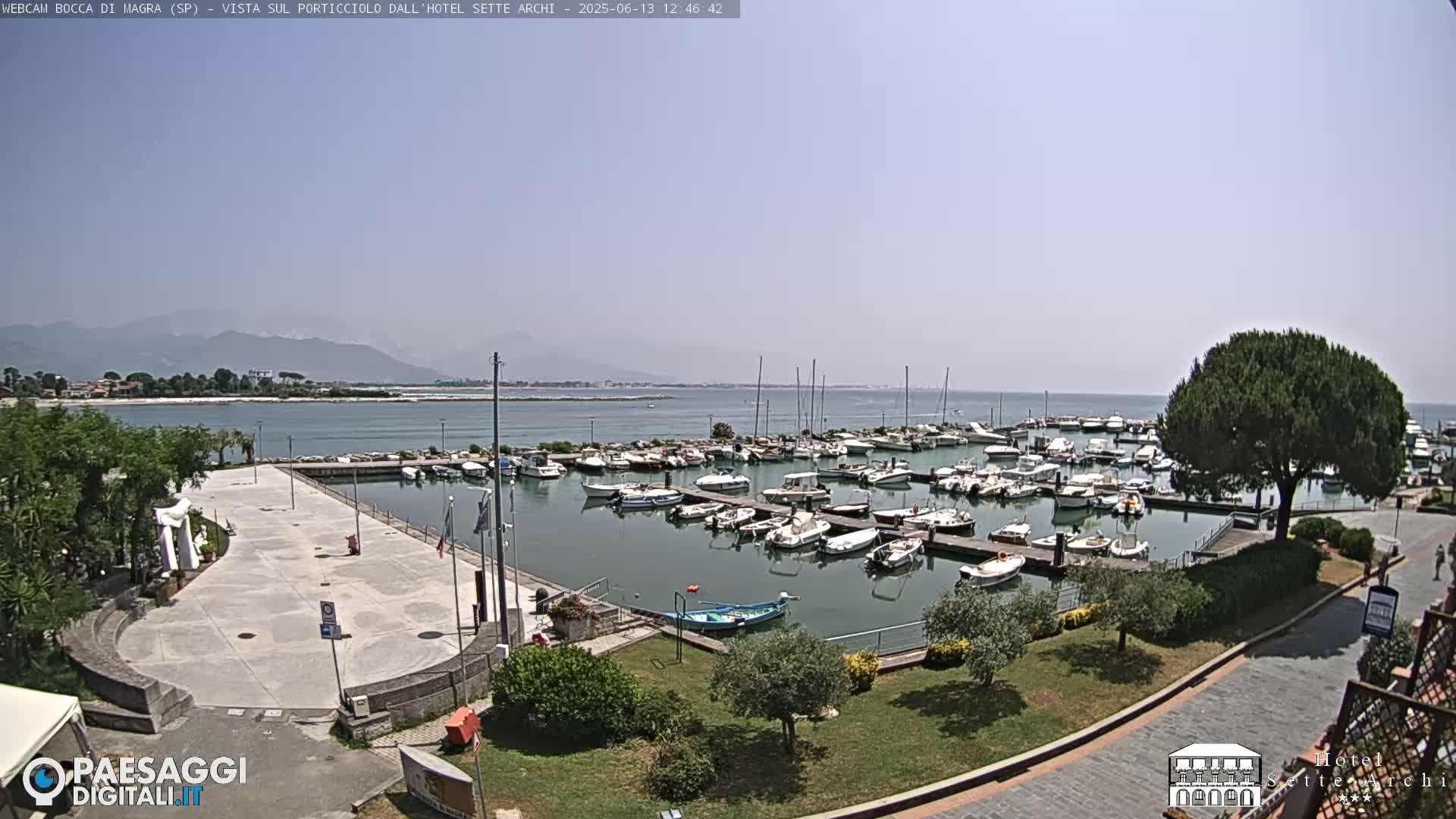 A marina filled with numerous boats is situated near a paved waterfront area, with mountains visible in the distance under a clear, sunny sky.