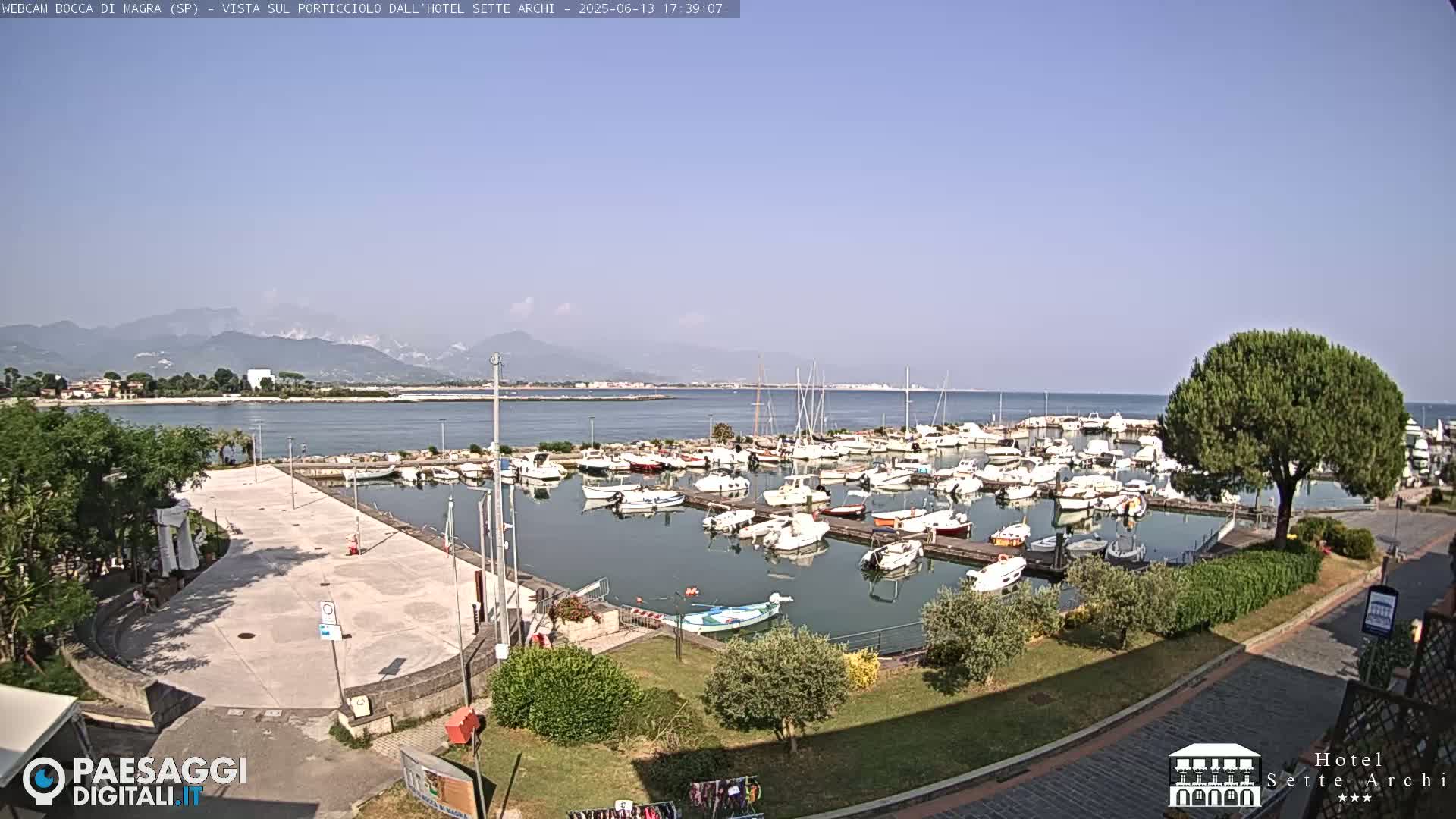 A marina filled with numerous boats is situated near a paved area and green landscaping, all overlooking a calm body of water and distant mountains under a clear sky.