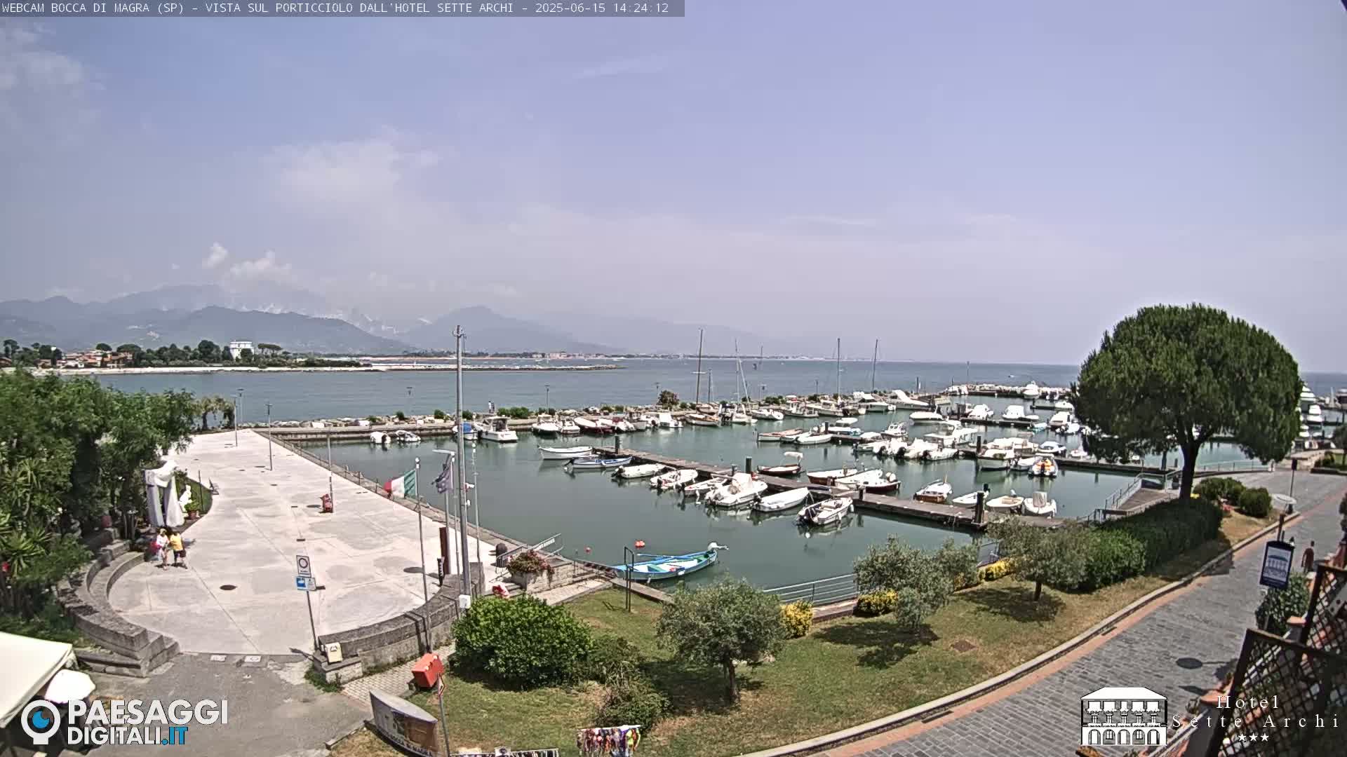 A sunny day shows a harbor filled with boats, a paved plaza, and mountains in the distance.