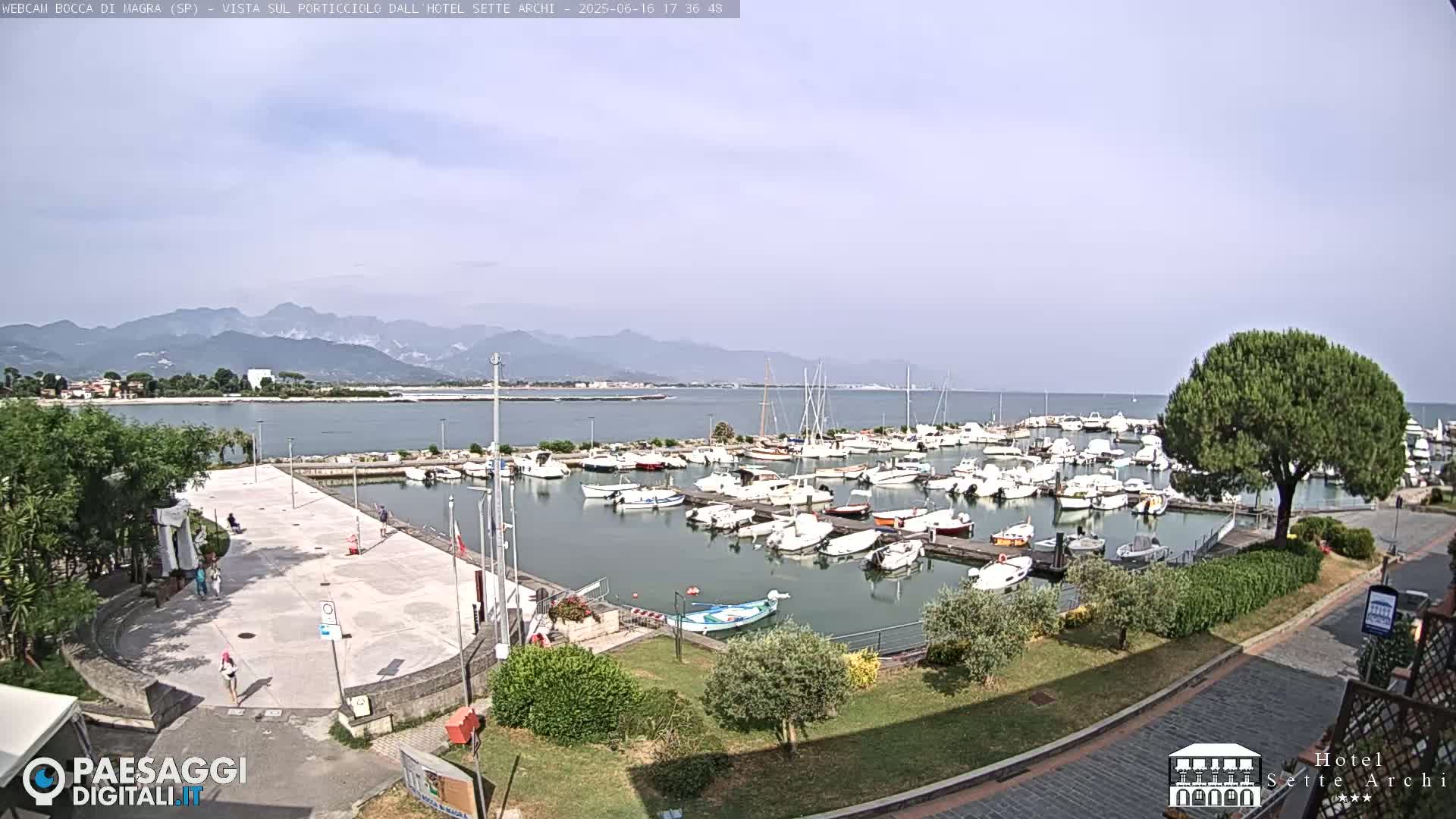 A marina filled with numerous boats is situated near a paved plaza, with a backdrop of mountains under a mostly clear sky.