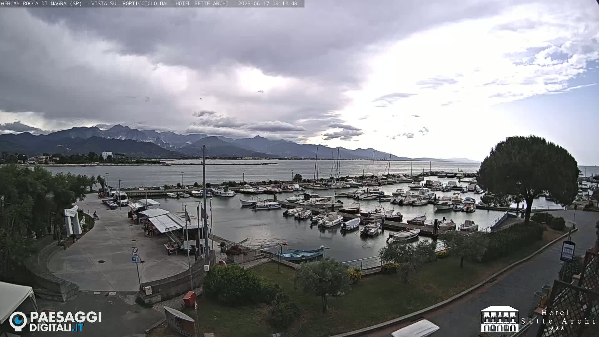 A harbor filled with numerous boats is situated near a shoreline, with mountains visible in the distance under a partly cloudy sky.