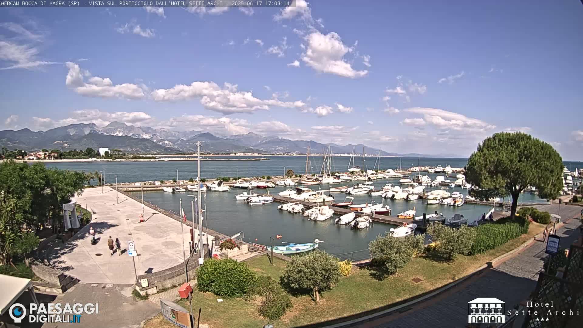A marina filled with boats is situated next to a paved area with a few people walking, all under a partly cloudy sky with mountains visible in the distance.