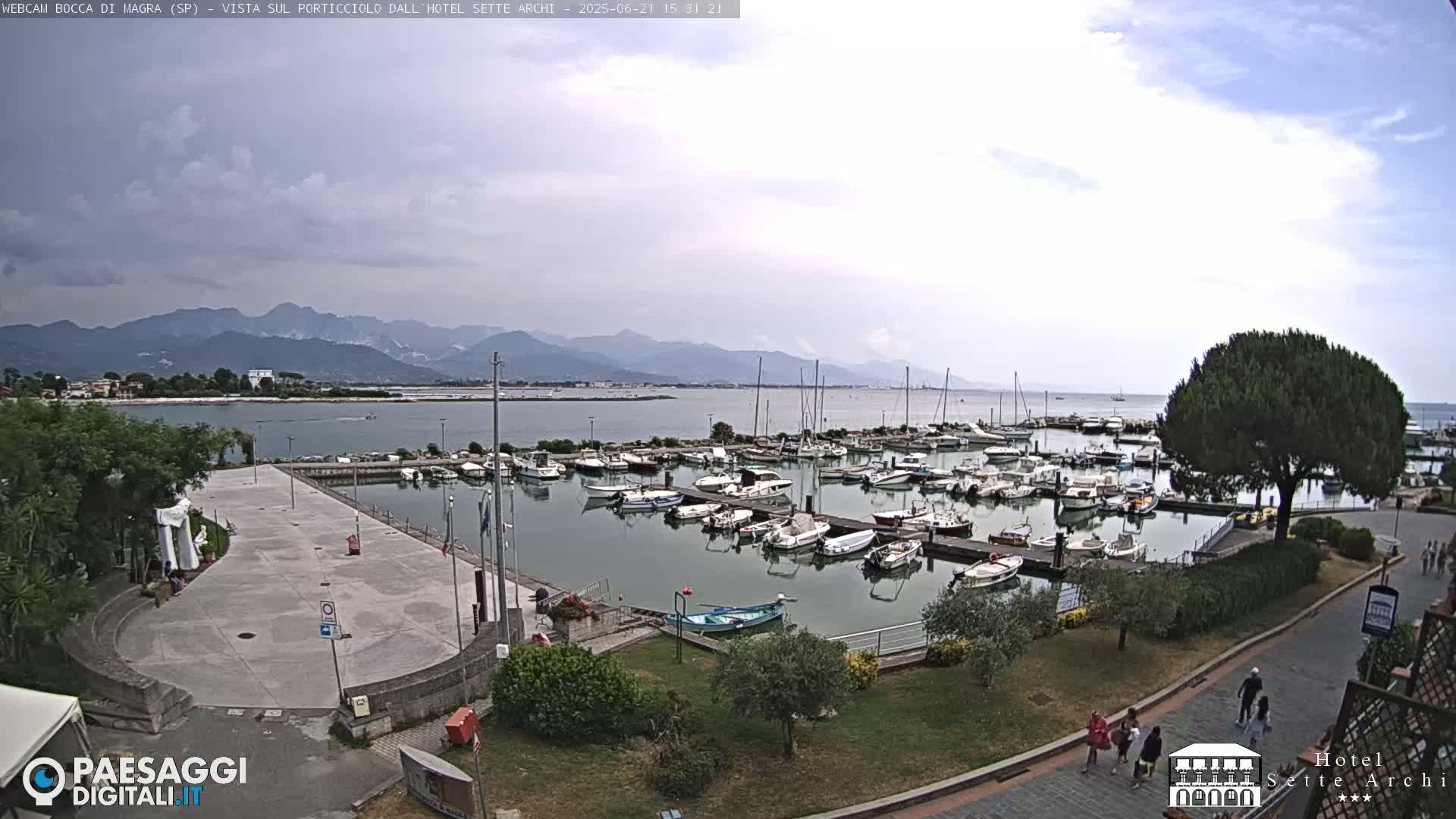 A harbor filled with numerous boats is situated near a paved area, with mountains visible in the distance under a partly cloudy sky.