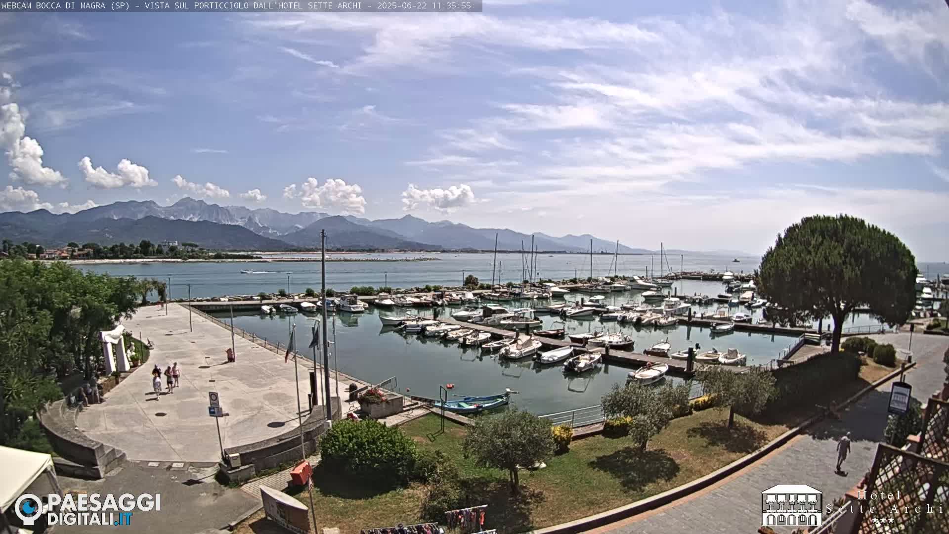 A marina filled with boats is situated near a paved walkway, with mountains visible in the background under a partly cloudy sky.
