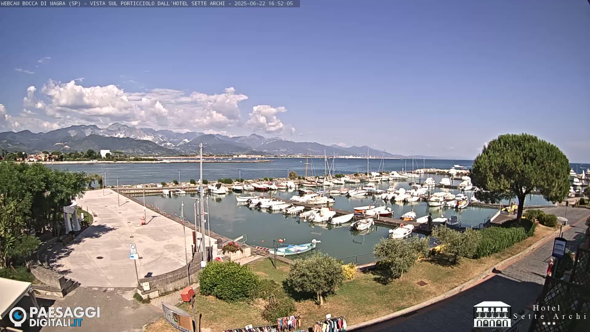 A marina filled with numerous boats is situated near a paved area, with mountains visible in the background under a partly cloudy sky.
