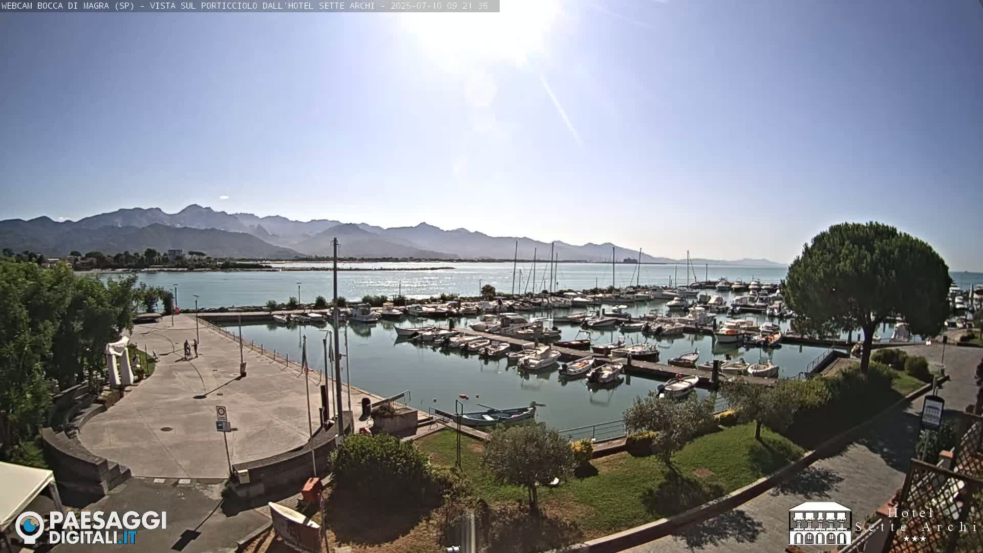 A marina filled with numerous boats is situated near a paved area, with mountains visible in the background under a clear, sunny sky.