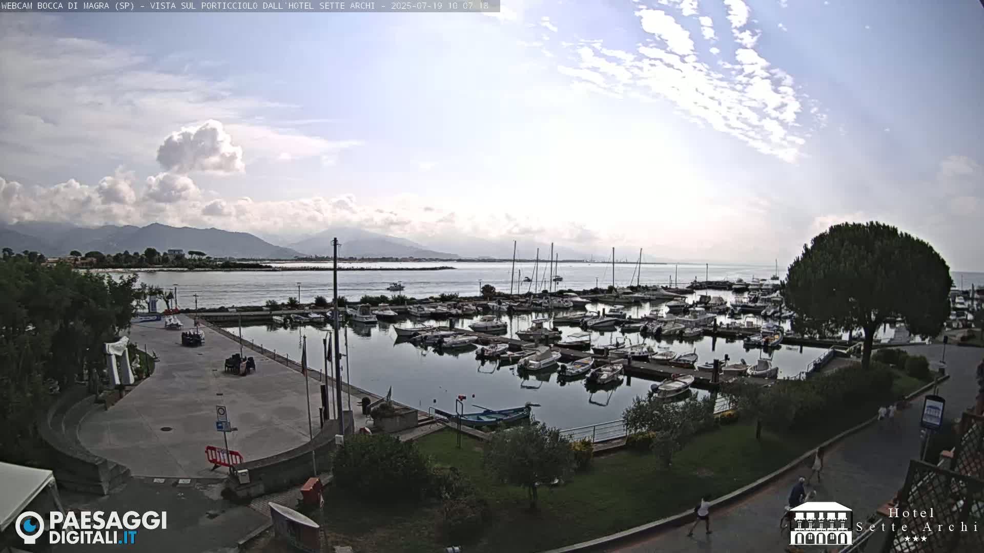 A partly cloudy day overlooks a marina filled with boats, a paved area, and green landscaping, with mountains in the distance.