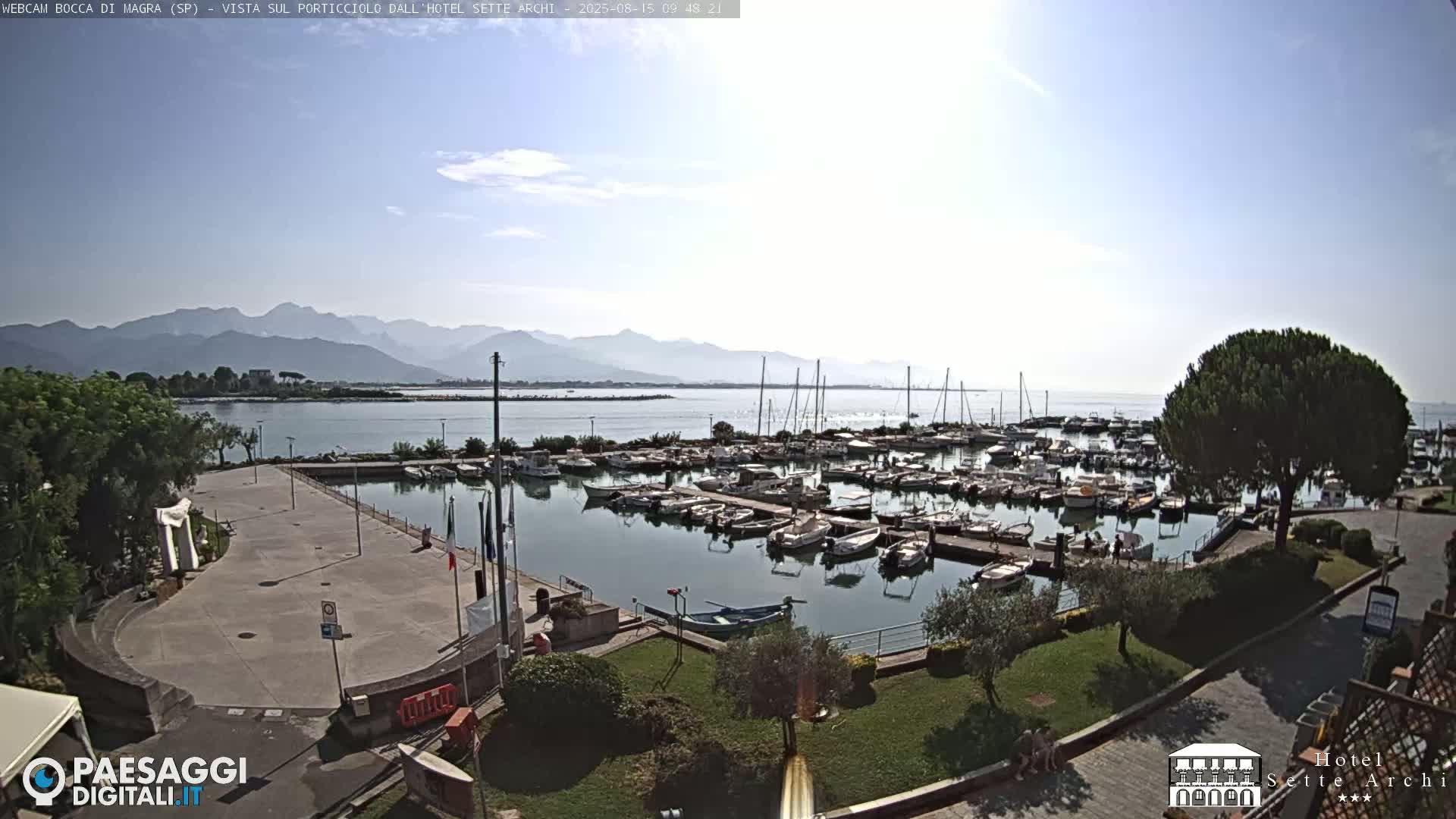A marina filled with boats is shown on a sunny day, with mountains in the distance.