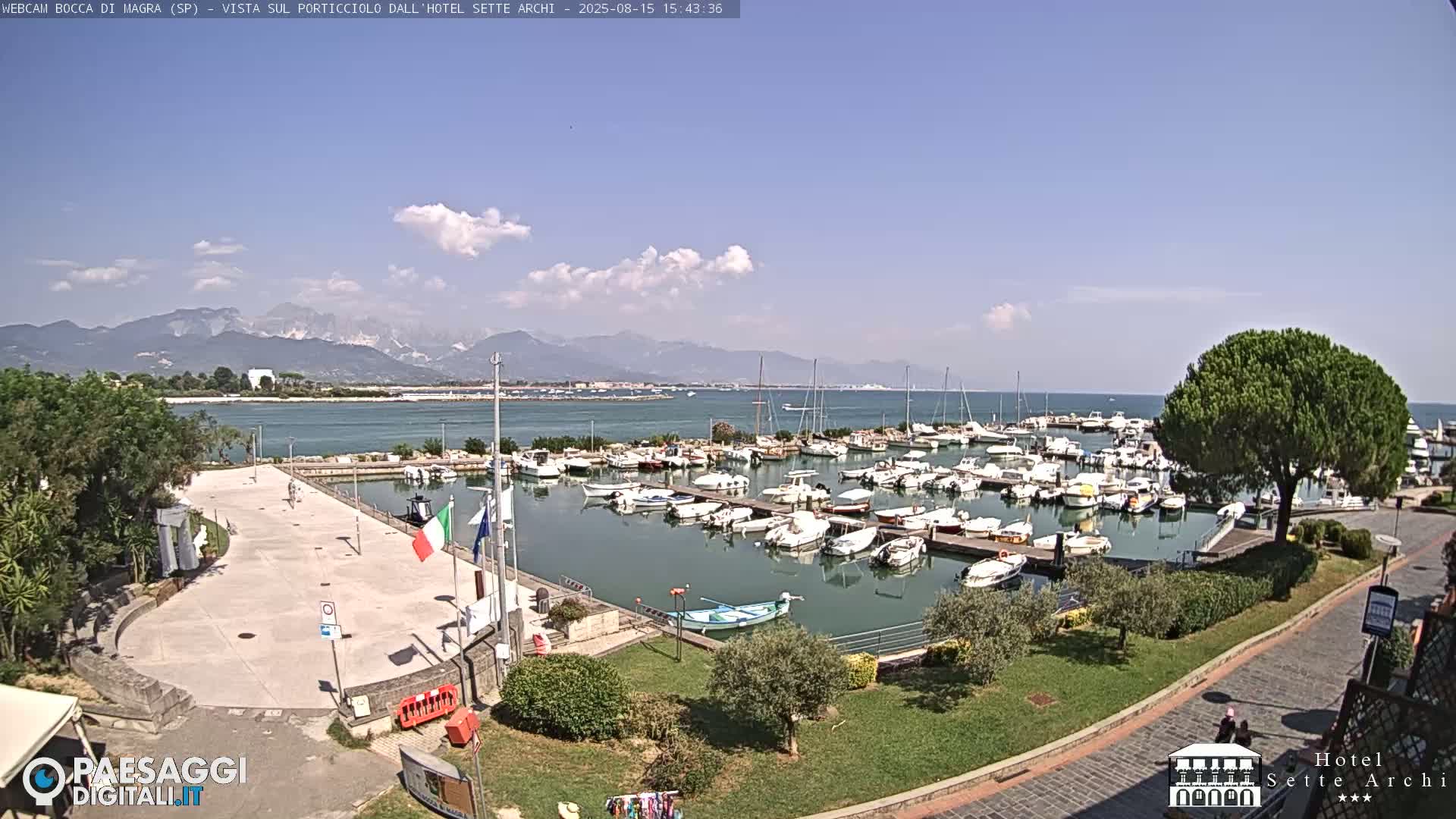 A marina filled with boats is shown on a sunny day, with mountains in the distance.
