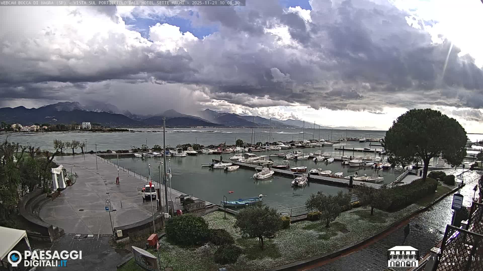 A marina filled with numerous boats is depicted under a dramatic, stormy sky with dark, heavy clouds and hints of sun breaking through, while the surrounding ground appears lightly covered in fresh hail, with distant mountains visible in the background.