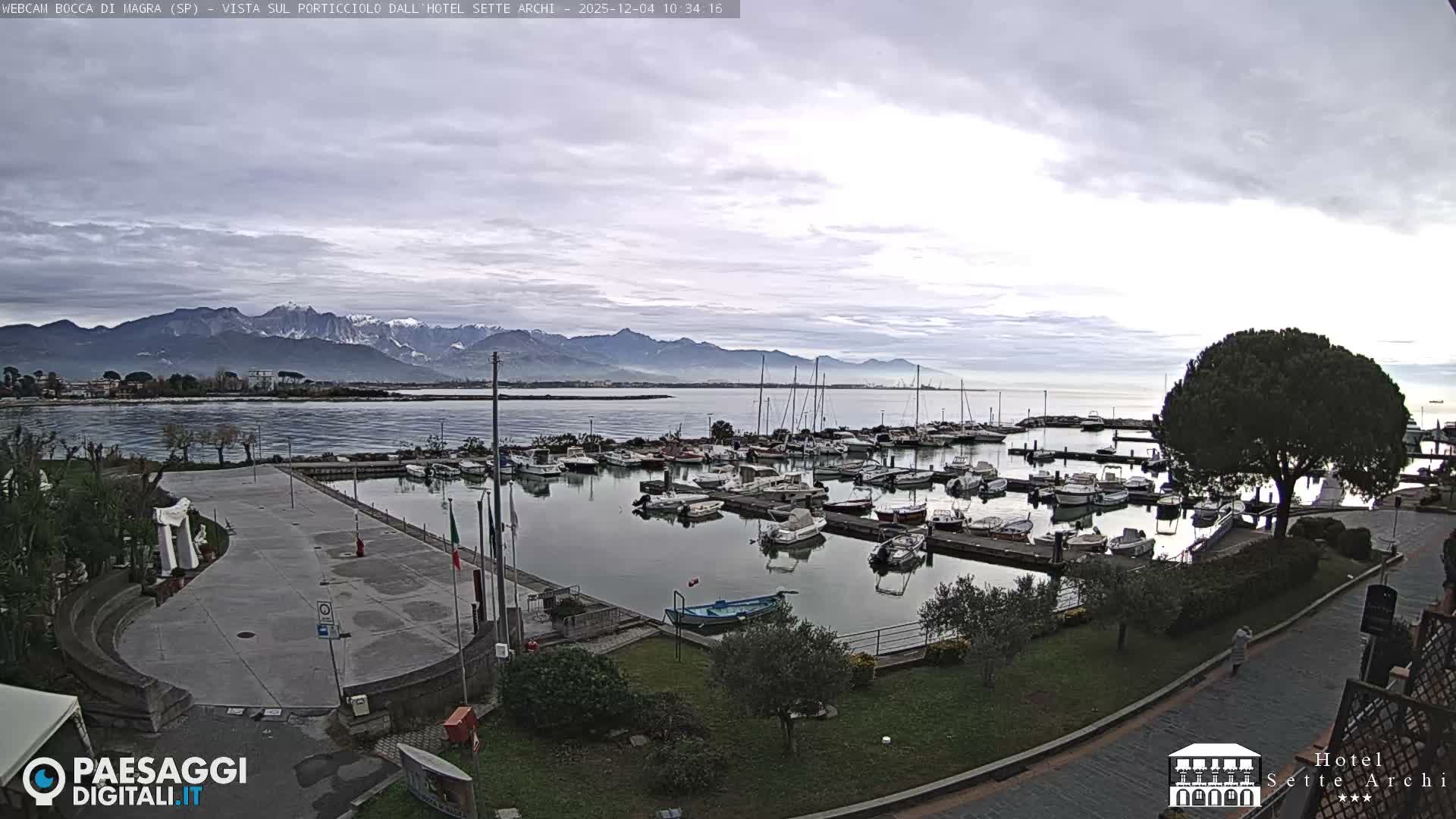 A wide view captures a bustling marina filled with boats on a calm body of water, flanked by a paved waterfront and distant, hazy mountains with some snow-capped peaks under an overcast sky.