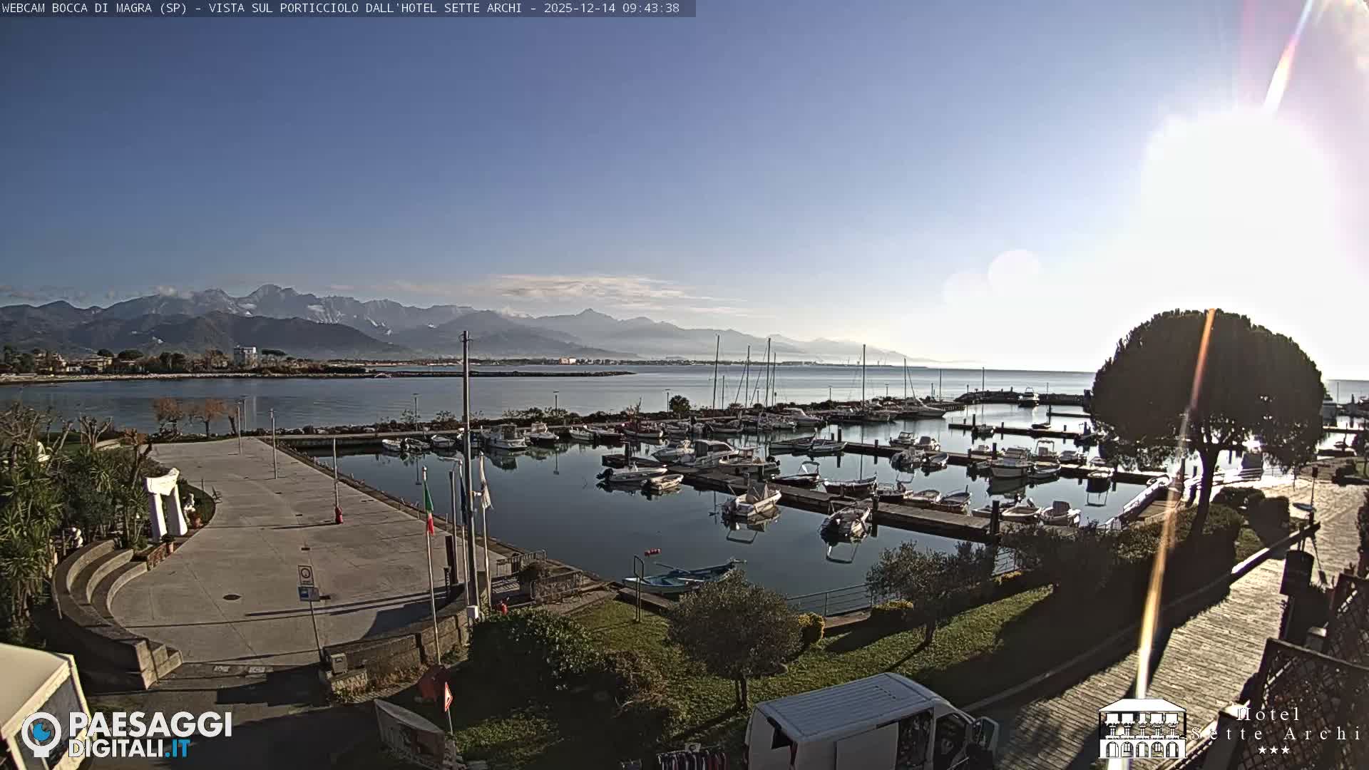 A wide view captures a bustling marina filled with boats on a calm body of water, flanked by a paved waterfront and distant, hazy mountains with some snow-capped peaks under an overcast sky.