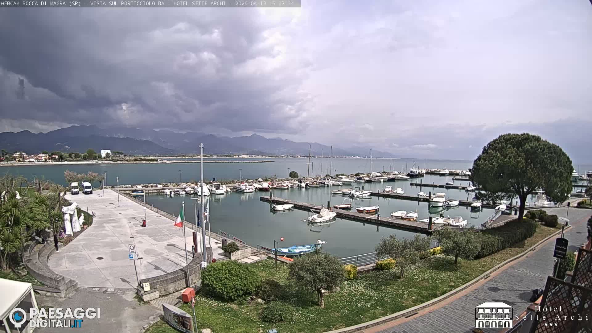 A wide view captures a bustling marina filled with boats on a calm body of water, flanked by a paved waterfront and distant, hazy mountains with some snow-capped peaks under an overcast sky.