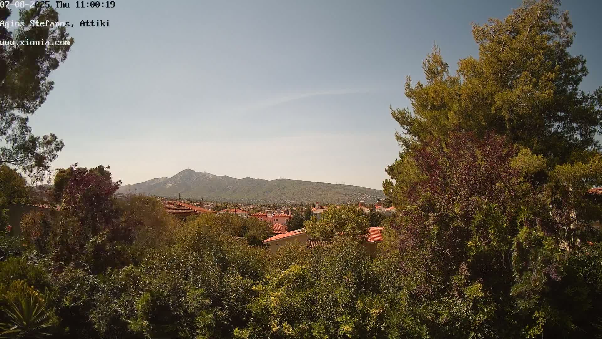 A view of a residential area with red-tiled roofs and lush green vegetation, under a clear sky, with mountains visible in the distance.