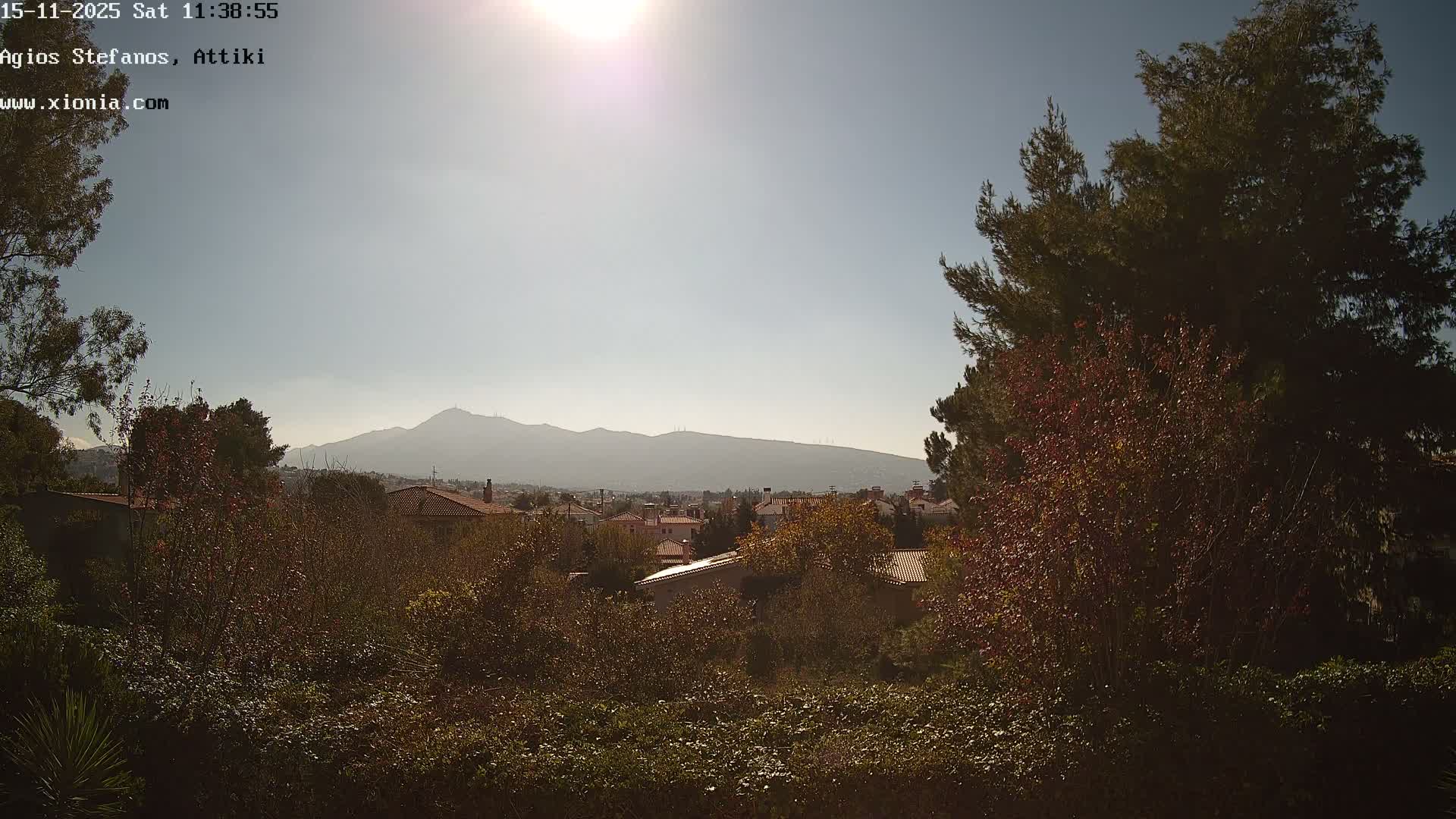 A sunny day with a clear blue sky overlooks a residential area with red-roofed houses surrounded by green and autumn-colored trees, all framed by hazy distant mountains.
