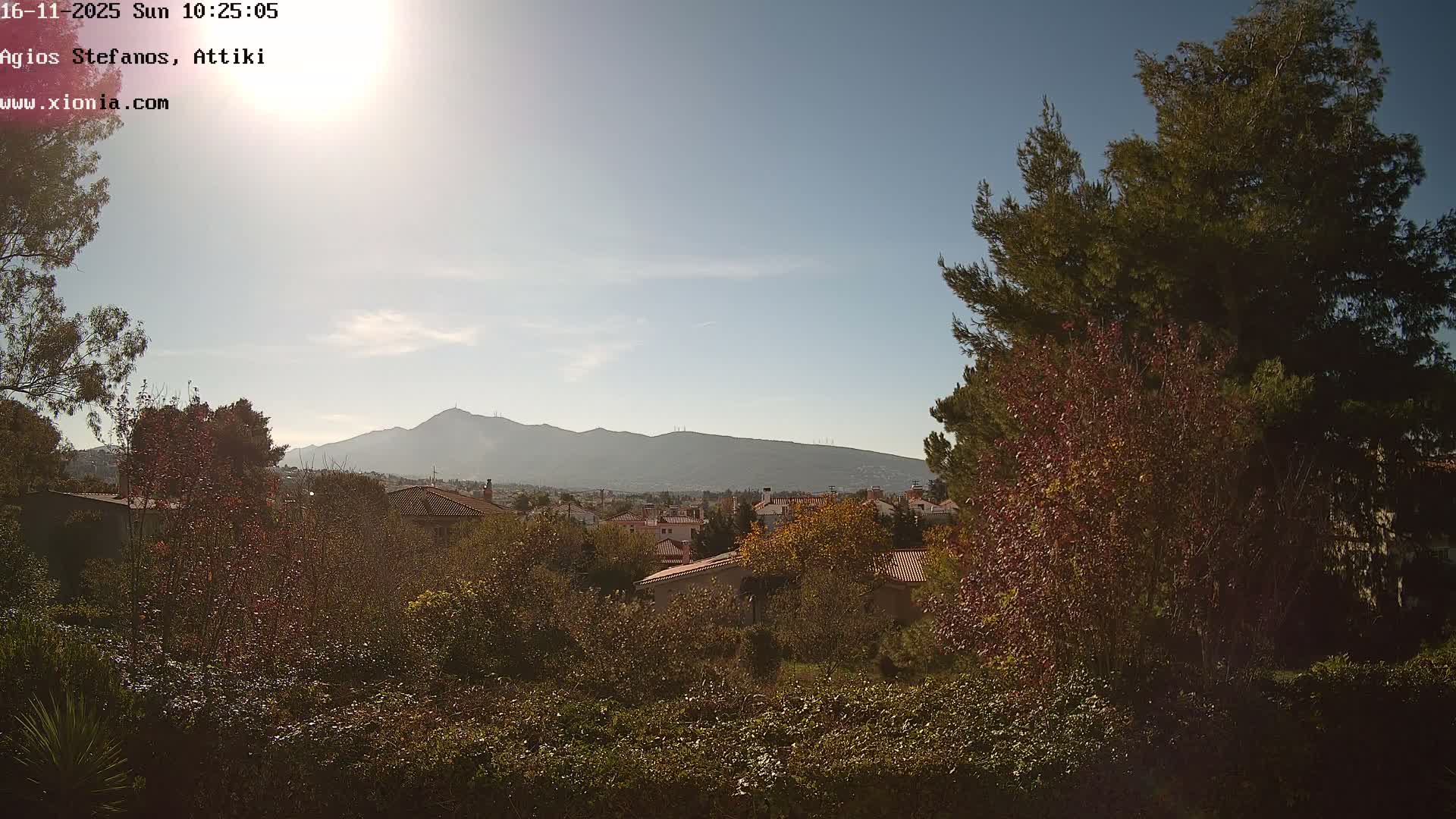The image shows a sunny, partially cloudy daytime view over a residential area with red-roofed houses nestled among trees displaying autumn colors and evergreens, all against a backdrop of distant mountains.