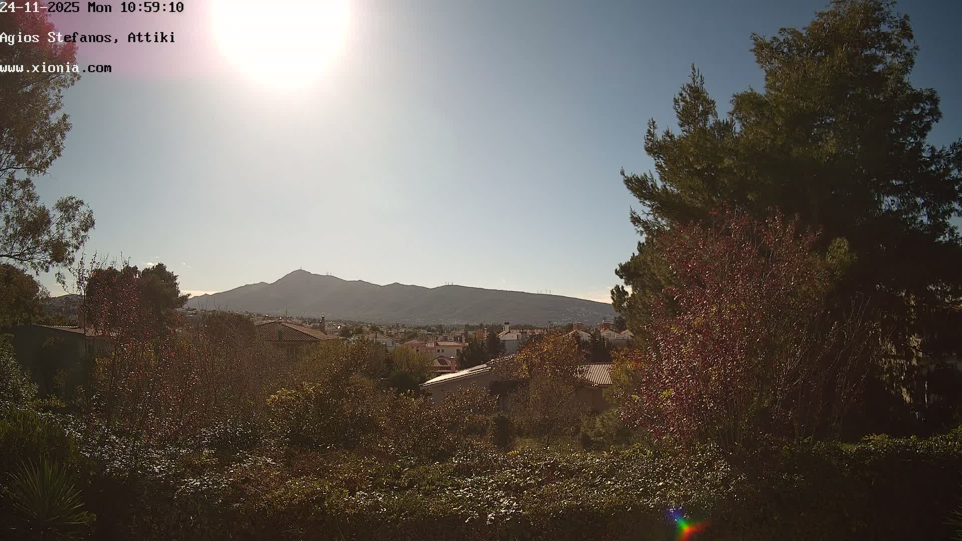 A panoramic view reveals a residential area with red-tiled roofs nestled among lush green and reddish autumn foliage, all set against a backdrop of distant mountains under a sunny and clear blue sky.