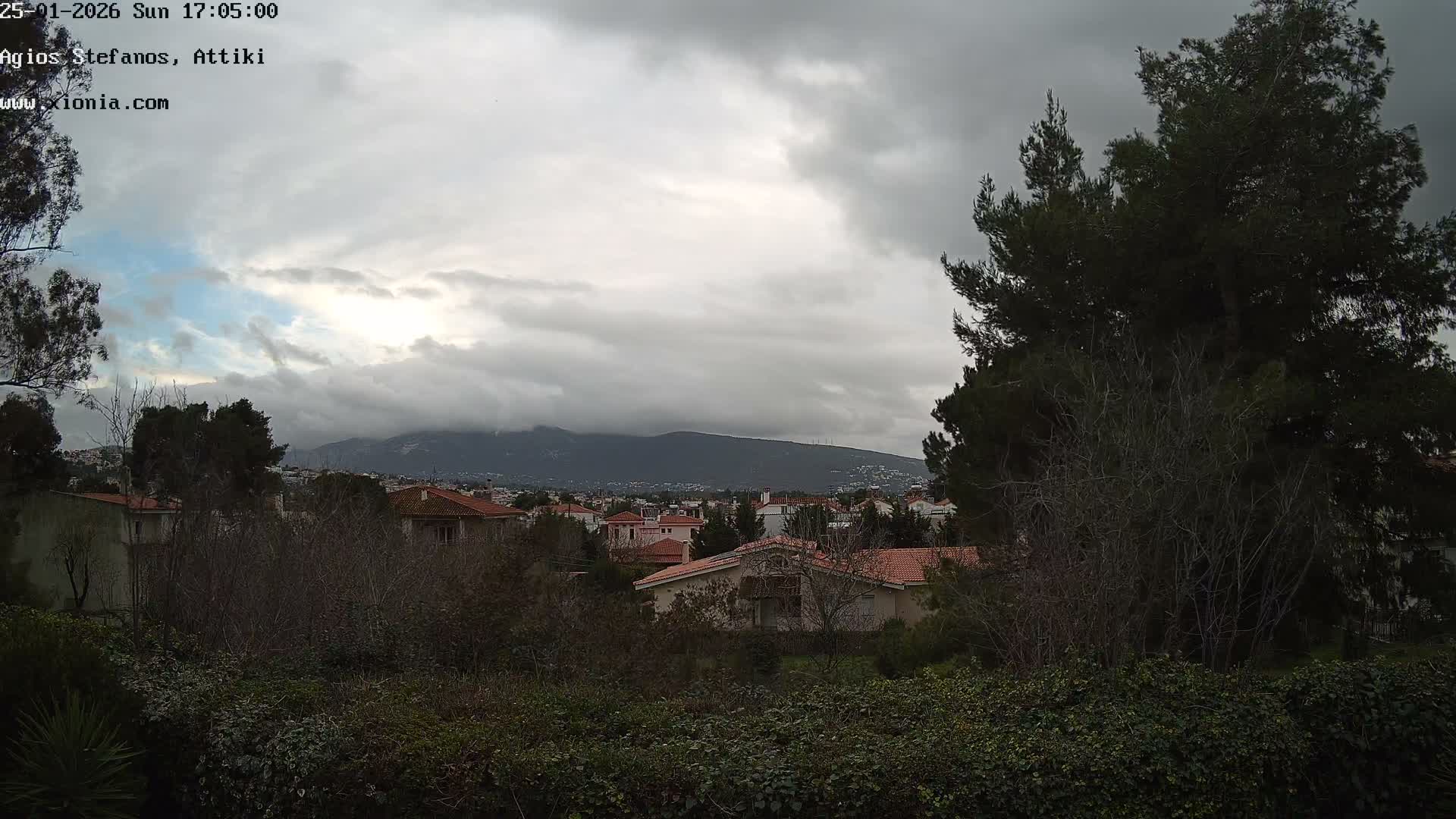 An outdoor scene depicts a residential area with red-tiled roofs nestled among trees and lush greenery, all under a predominantly overcast sky with a glimpse of blue, and distant mountains shrouded in clouds.