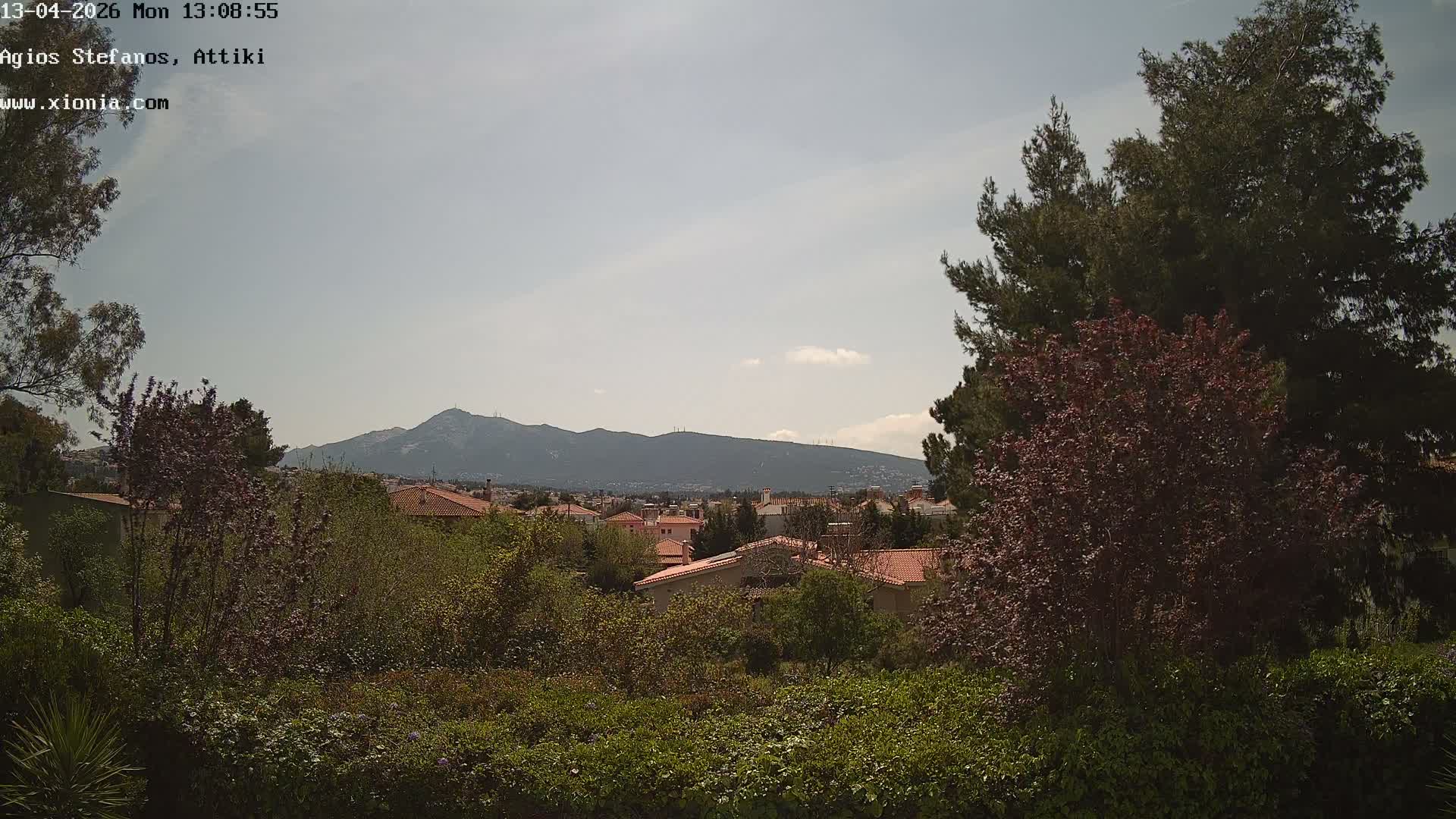 An outdoor scene depicts a residential area with red-tiled roofs nestled among trees and lush greenery, all under a predominantly overcast sky with a glimpse of blue, and distant mountains shrouded in clouds.