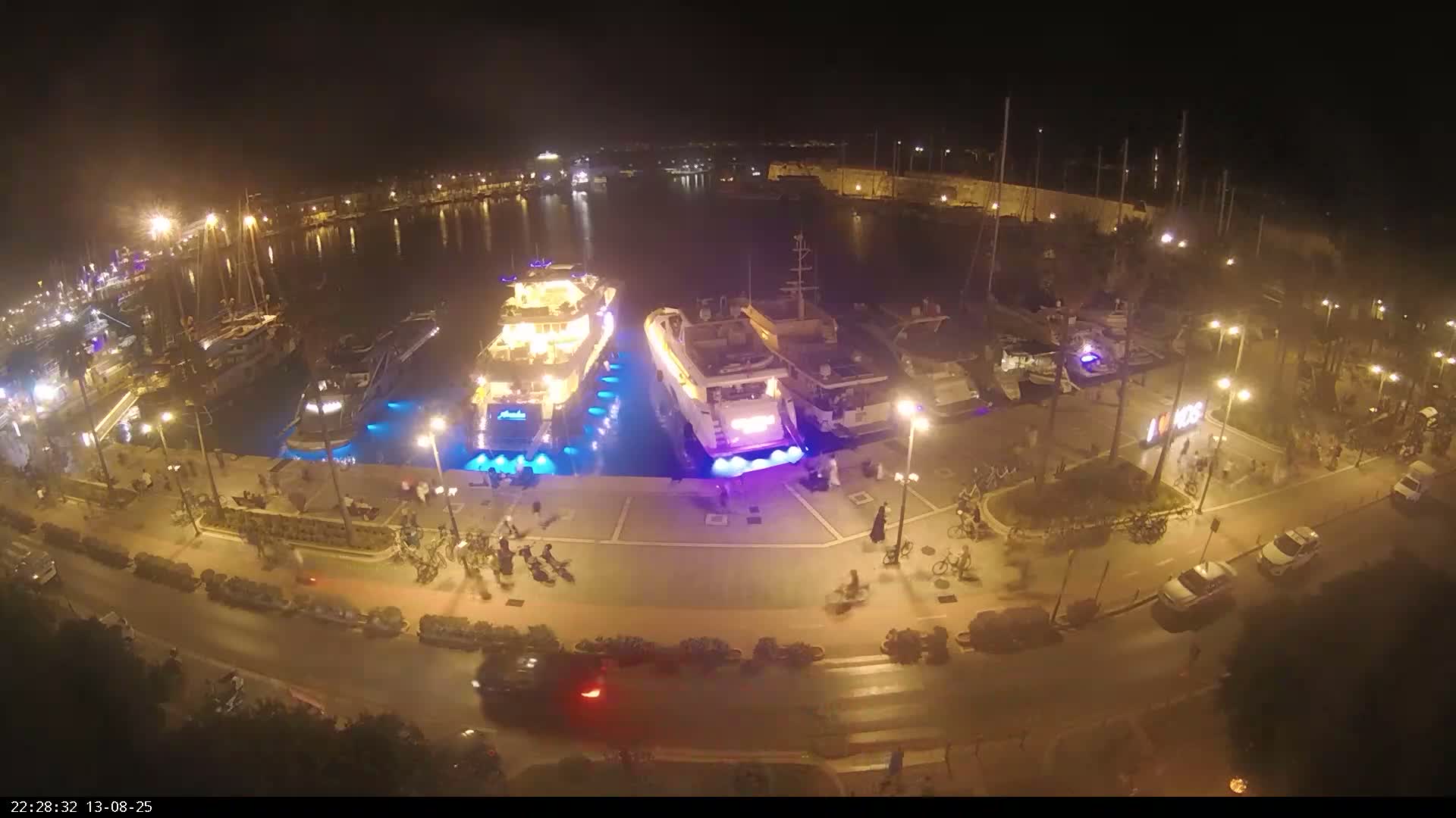 A nighttime aerial view shows a harbor filled with boats and yachts illuminated by lights, with people walking and biking along a nearby roadway.