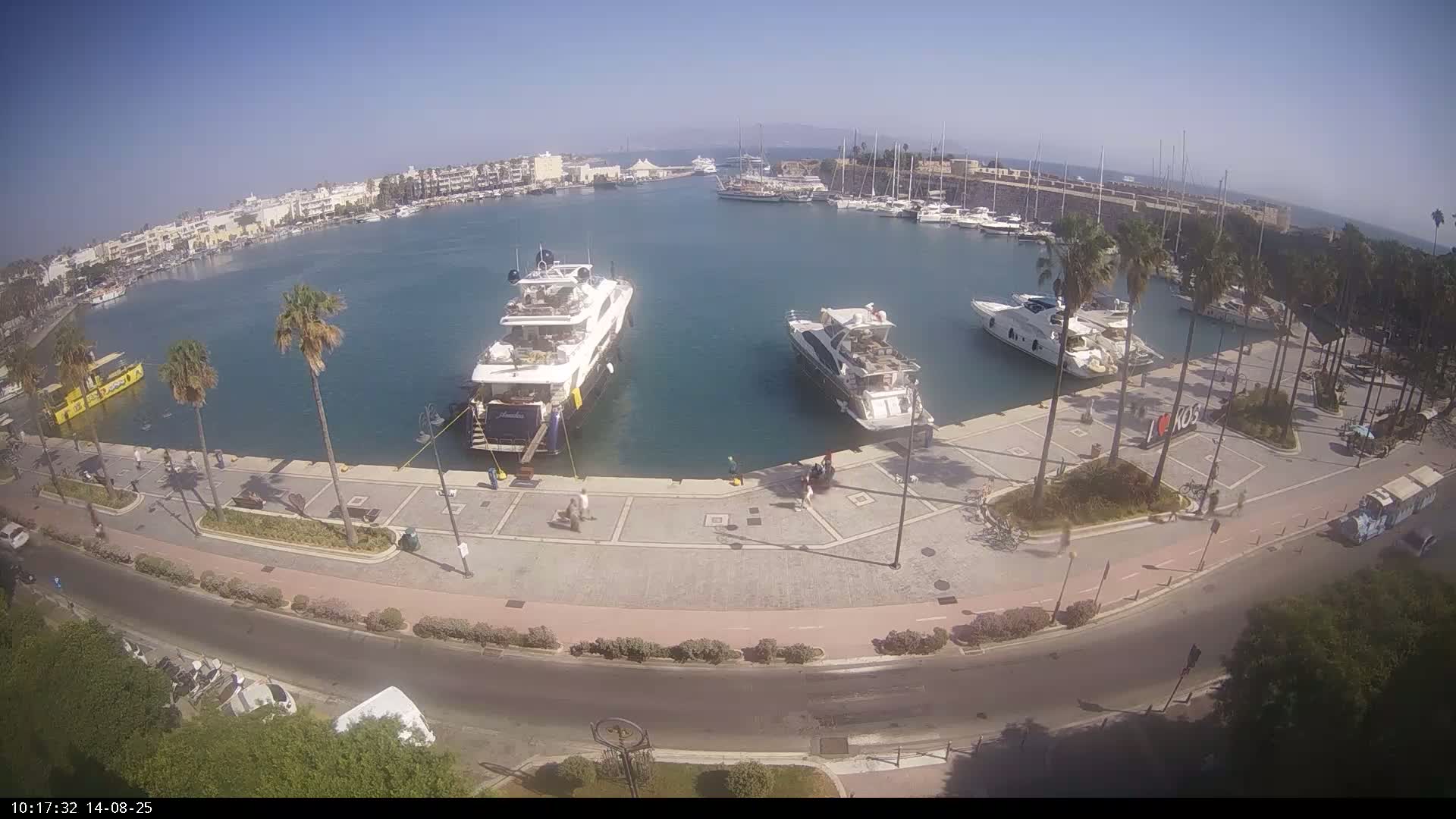 A sunny day at a marina shows several yachts and boats docked, with palm trees and people walking along a paved waterfront path.