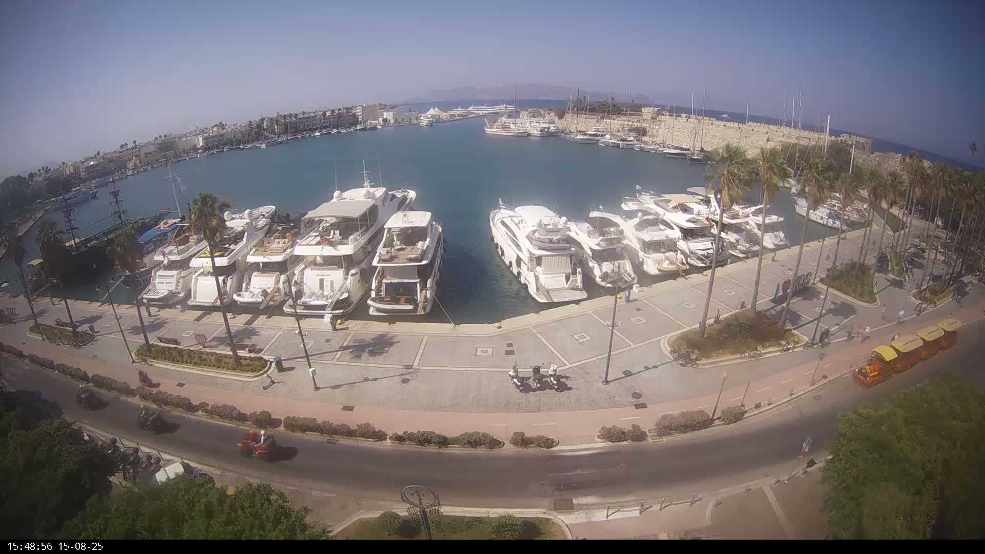A sunny day at a marina shows several yachts and boats docked, with palm trees and people walking along a paved waterfront path.