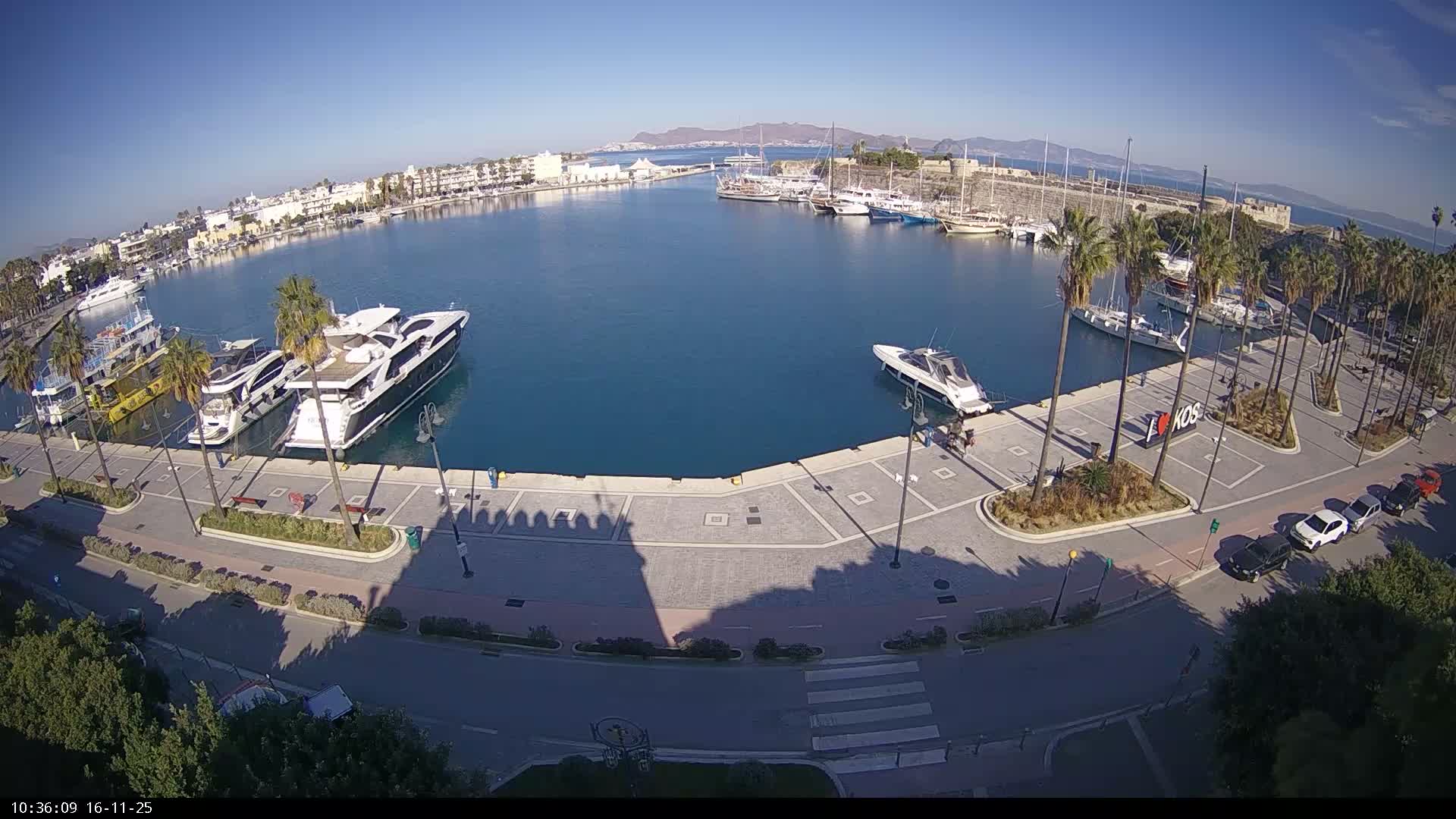 An aerial view shows a sunny day over a bustling harbor filled with yachts and various boats, flanked by a city with palm trees, a waterfront promenade, roads with parked cars, and a historical fortress, with mountains visible in the distance across the calm blue water.