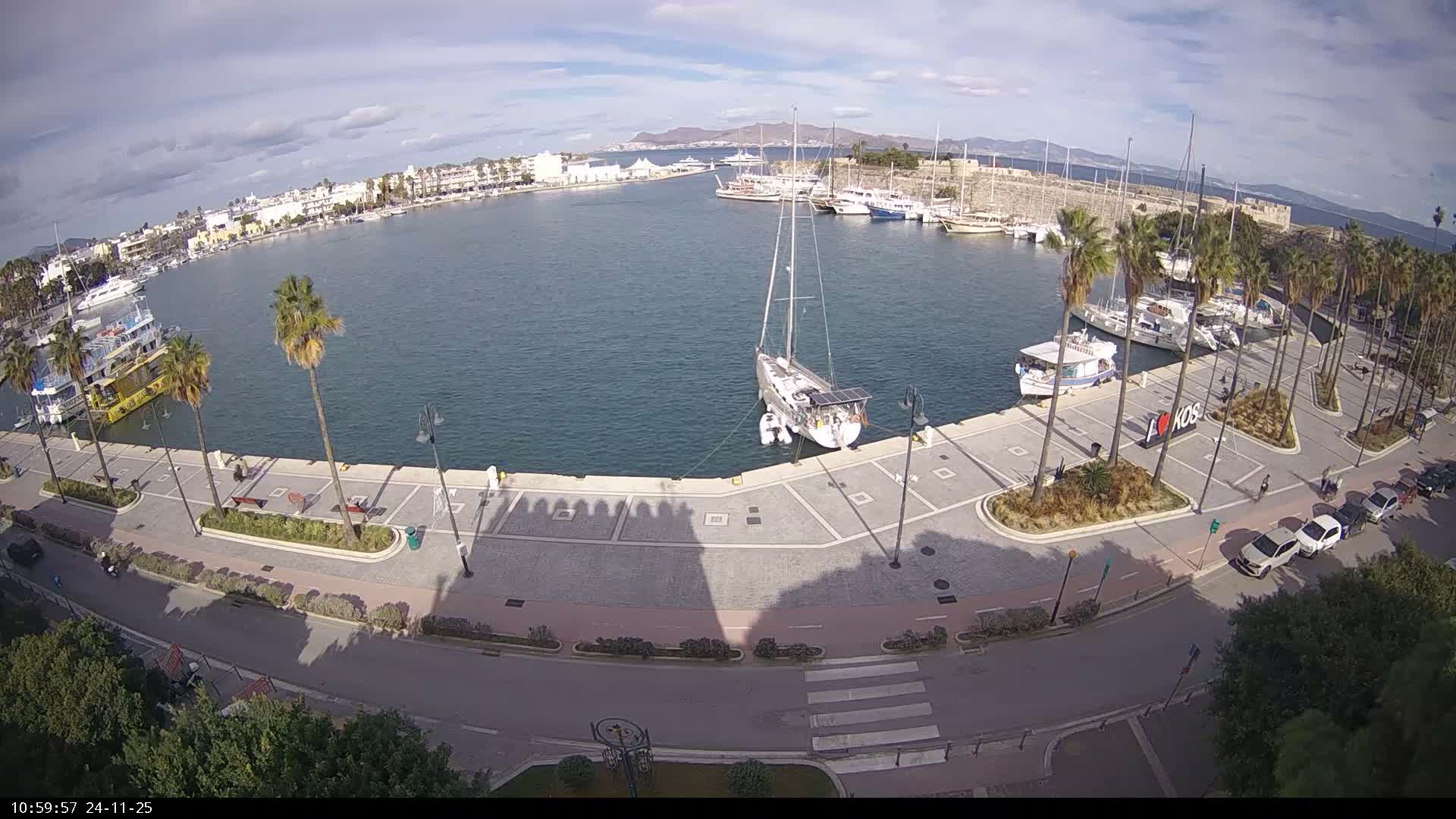 A wide aerial view captures a sunny Mediterranean harbor on a partly cloudy day, featuring numerous yachts and boats docked along a palm-lined promenade, with a town, a historic fortified wall, and distant mountains forming the backdrop.