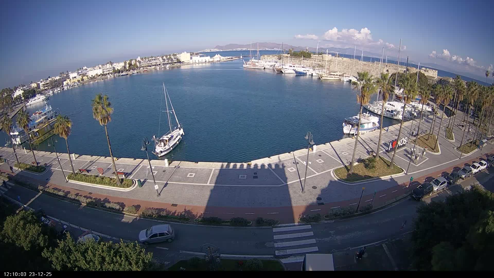 An aerial view reveals a sunny harbor with numerous boats docked along its edge and one sailboat in the center, encircled by a palm-lined promenade, coastal buildings, a road with cars, and a historic stone fort wall, all under a clear blue sky with a few scattered clouds.