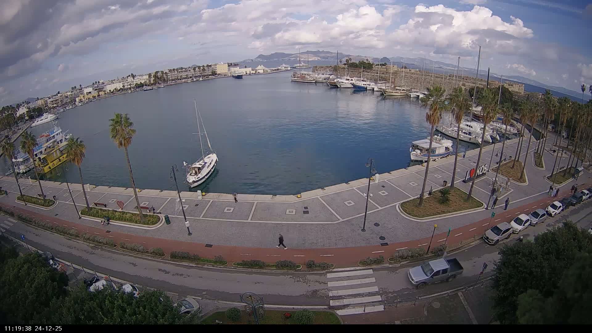 An aerial view reveals a sunny harbor with numerous boats docked along its edge and one sailboat in the center, encircled by a palm-lined promenade, coastal buildings, a road with cars, and a historic stone fort wall, all under a clear blue sky with a few scattered clouds.