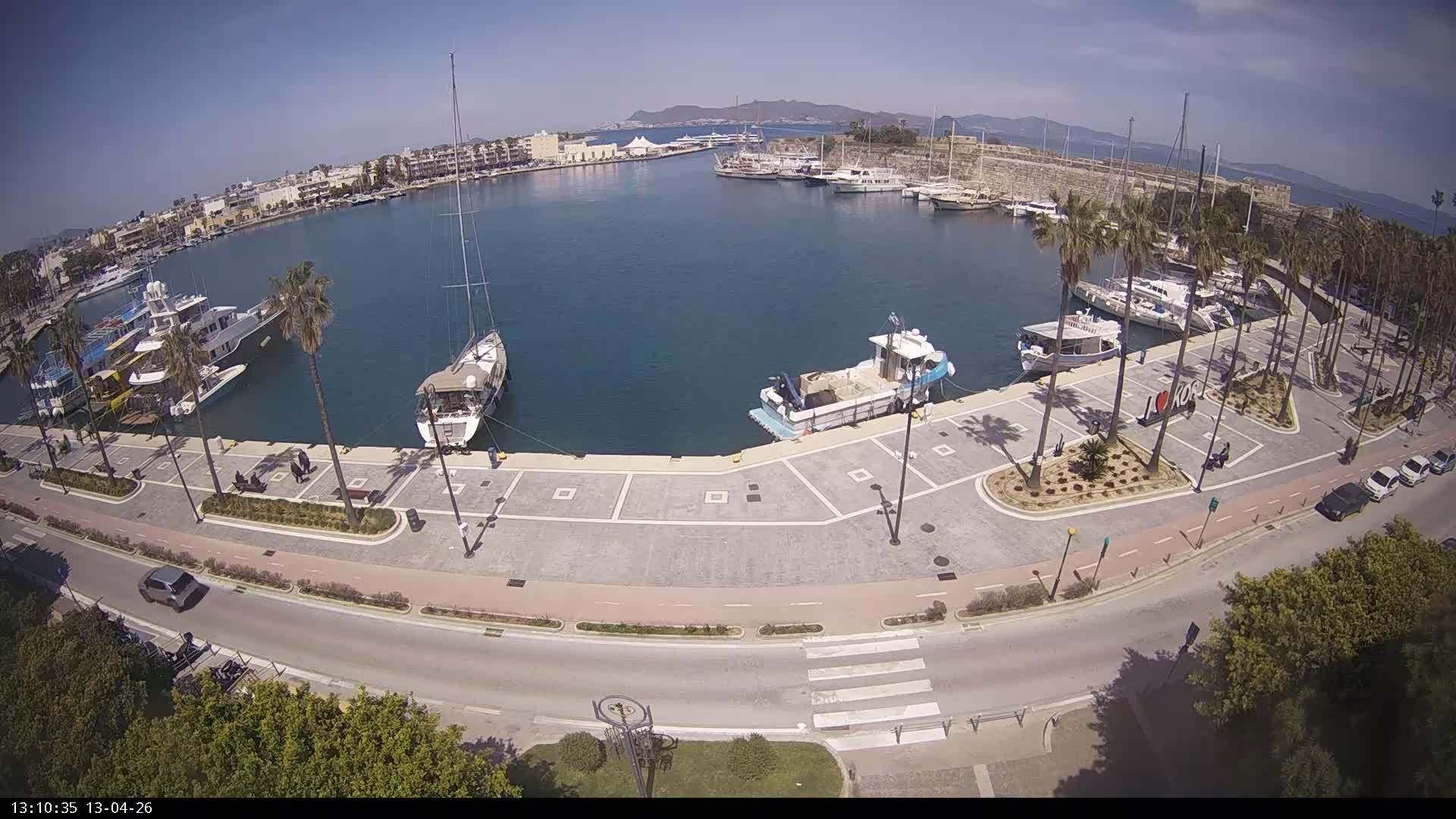 An overcast sky looms over a bustling harbor filled with numerous boats, bordered by a palm-lined promenade, a road with cars, and surrounding buildings, with distant mountains visible.