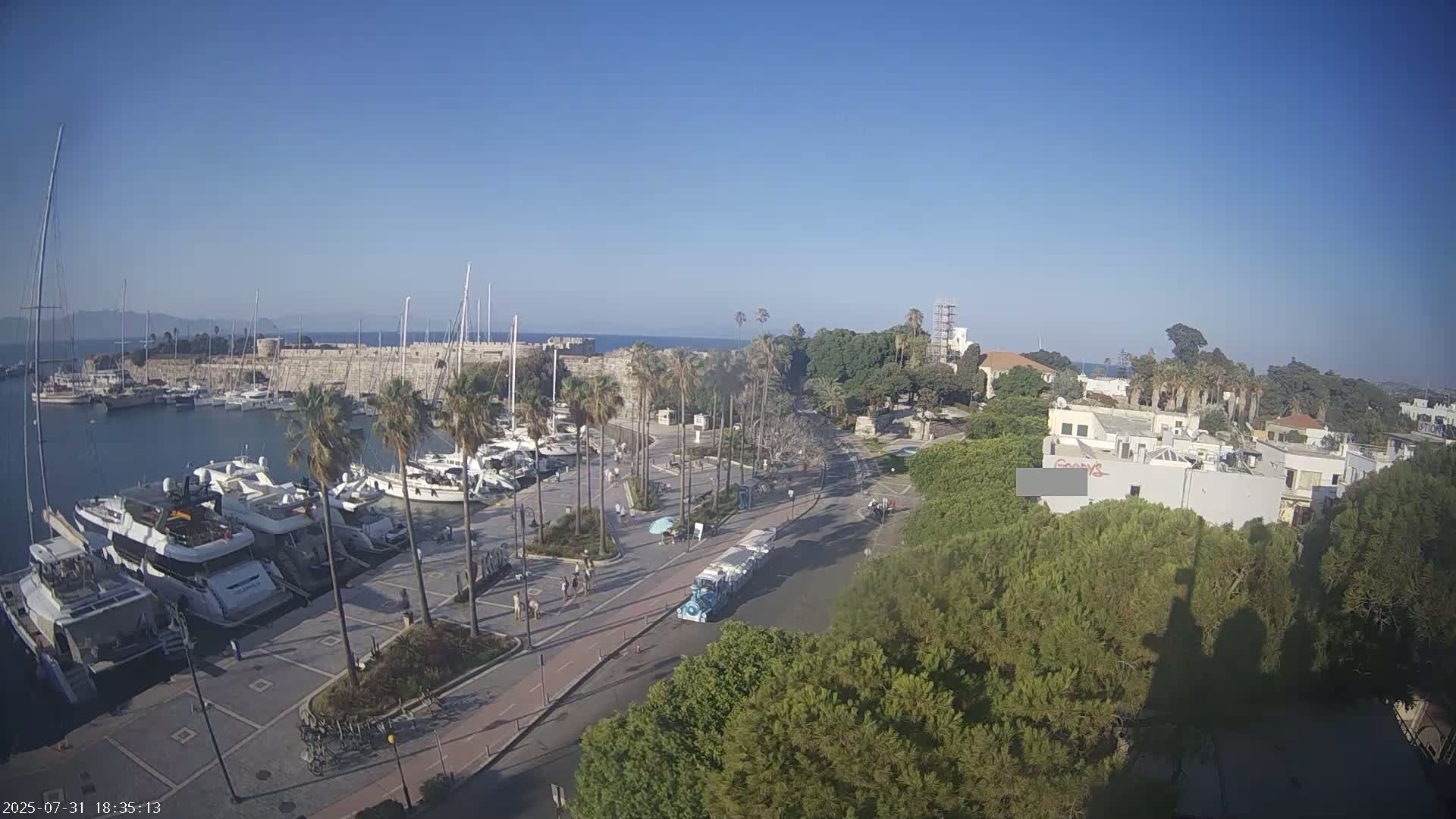 A harbor filled with yachts and boats is situated next to a stone wall, with palm trees lining a paved area and a tourist train driving along a road, all under a clear blue sky.