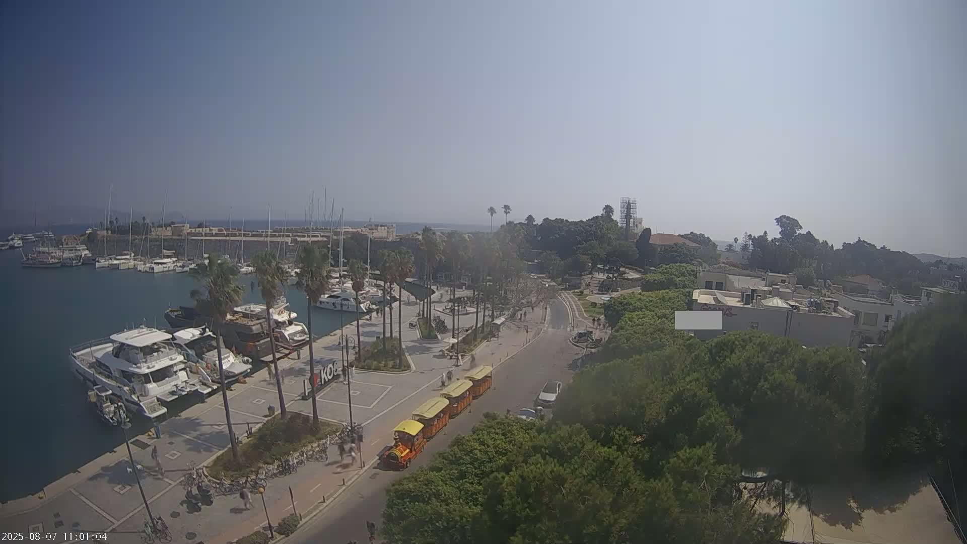 A marina filled with yachts is visible next to a road with a train of yellow tourist cars, under a clear, sunny sky.